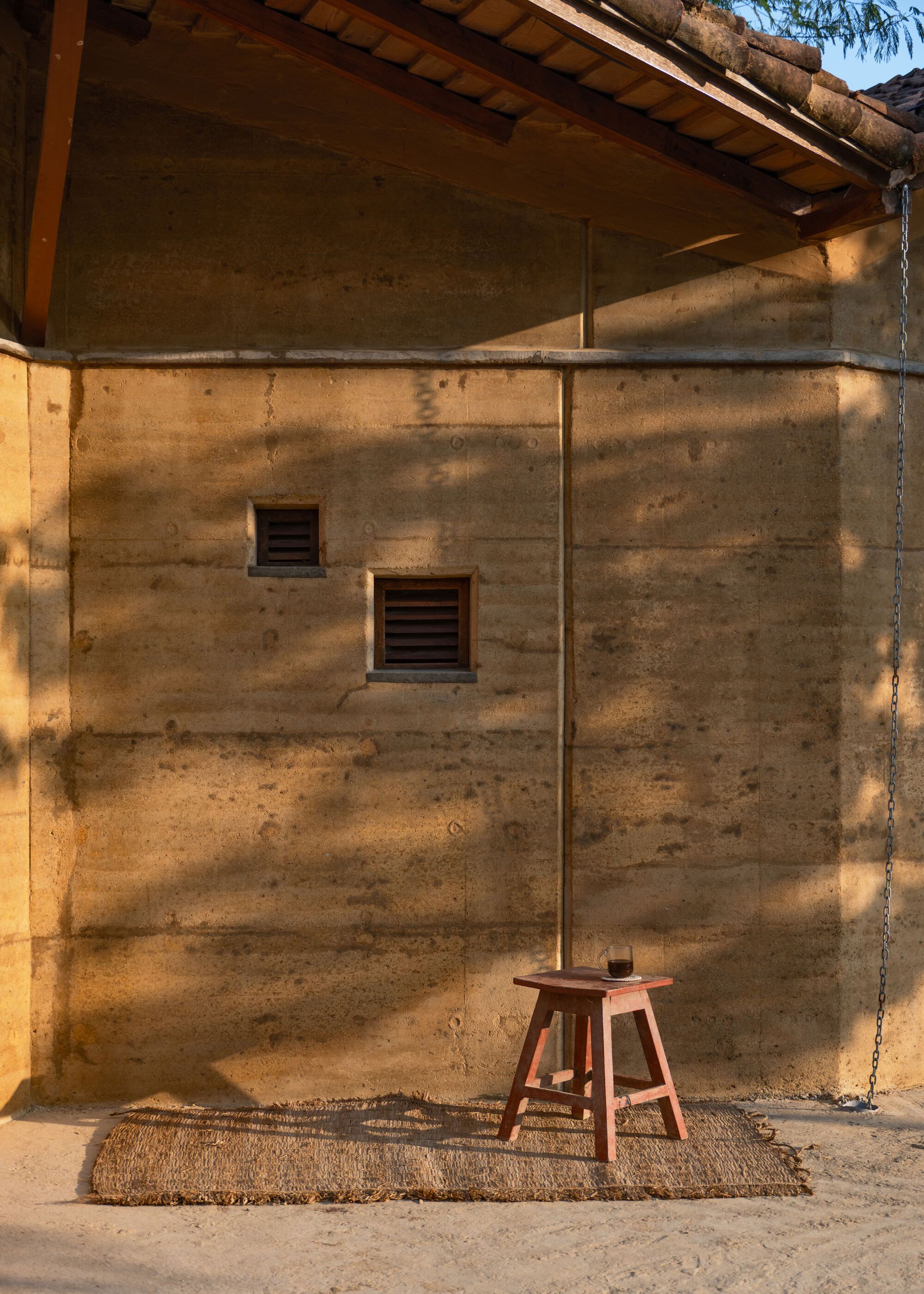 Layered rammed earth wall with two small louvred vents, a wooden stool on a woven mat and a hanging chain in afternoon shade