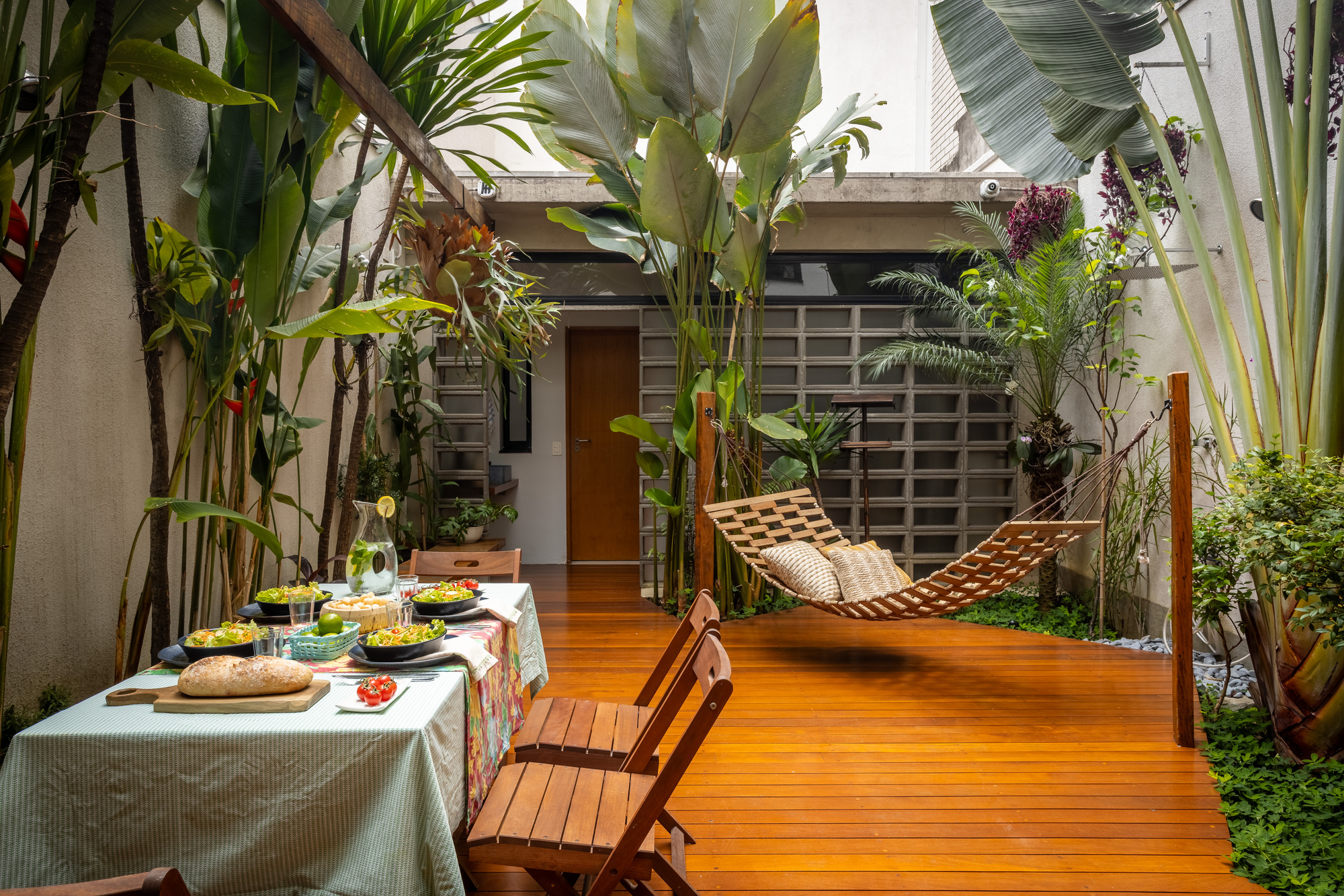 Timber-decked courtyard with a slatted hammock, set dining table and tropical plants in front of a glass block wall
