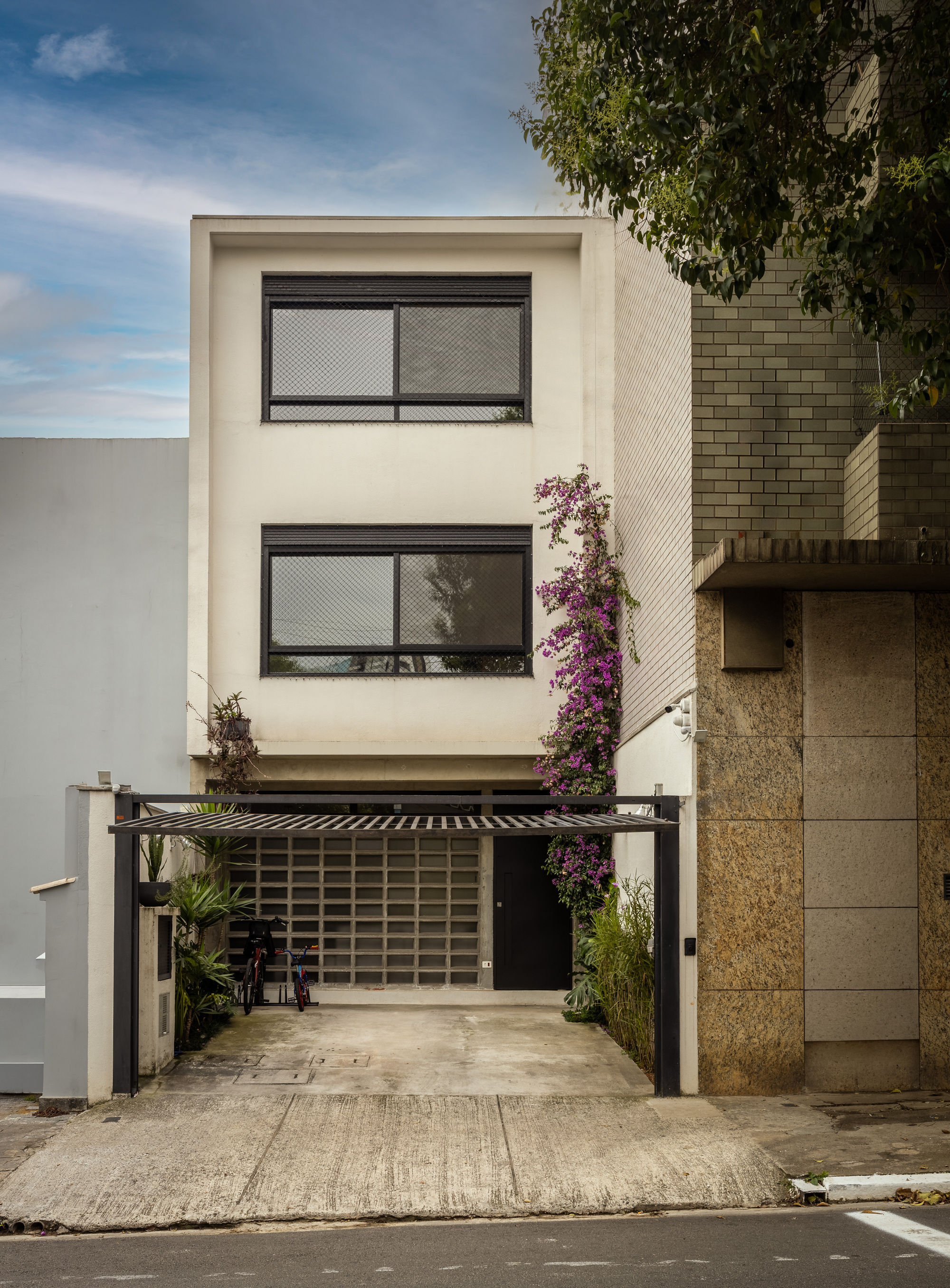 Street facade of a narrow three-storey house with black-framed windows, a steel carport canopy and flowering bougainvillea under a cloudy sky