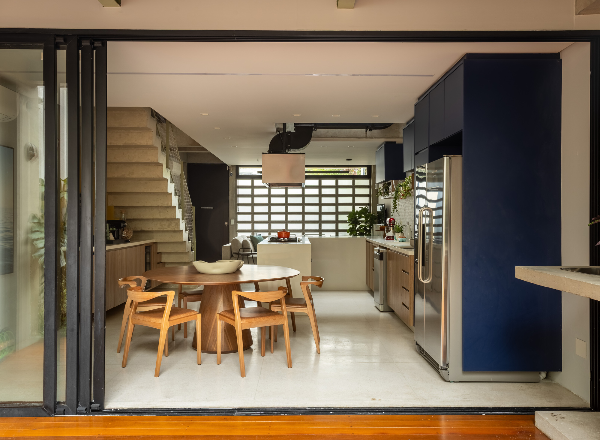 View through open folding glass doors to a round walnut dining table, navy kitchen cabinets and a concrete stair