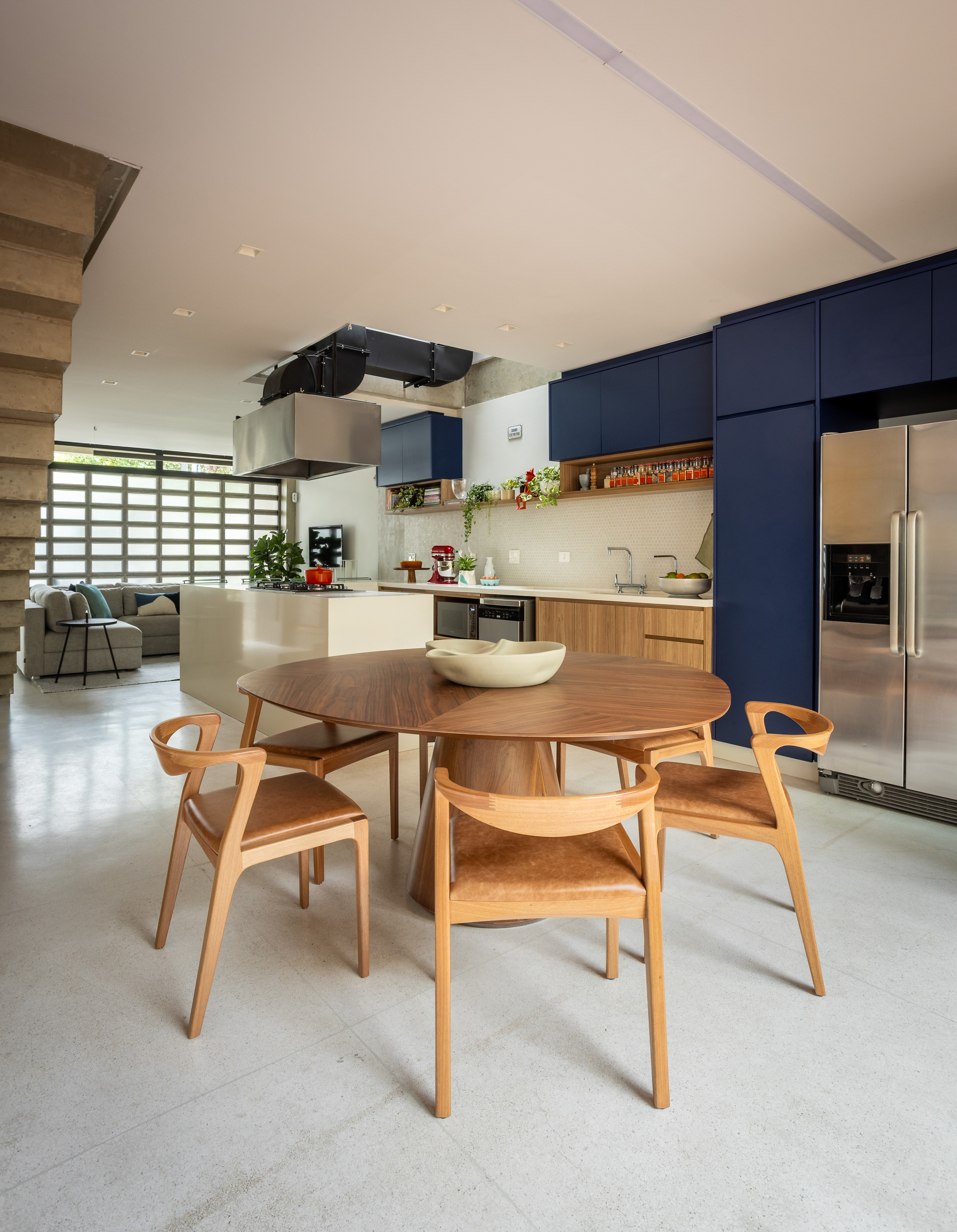 Round wooden dining table with leather-seated chairs in front of the navy kitchen, with a stepped concrete stair at left