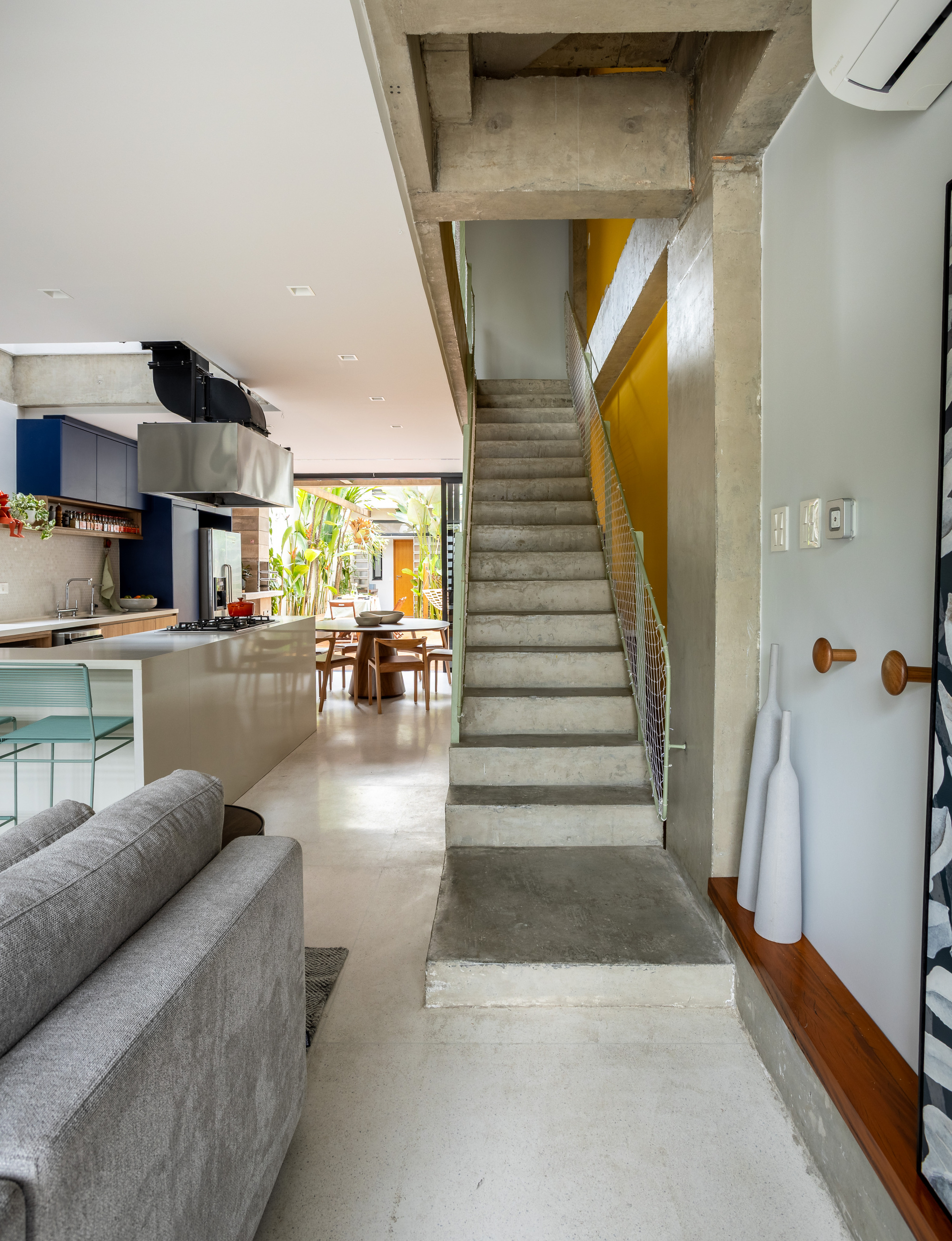Exposed concrete staircase with a mesh guard rising beside the kitchen island, with a courtyard glimpsed through glass beyond