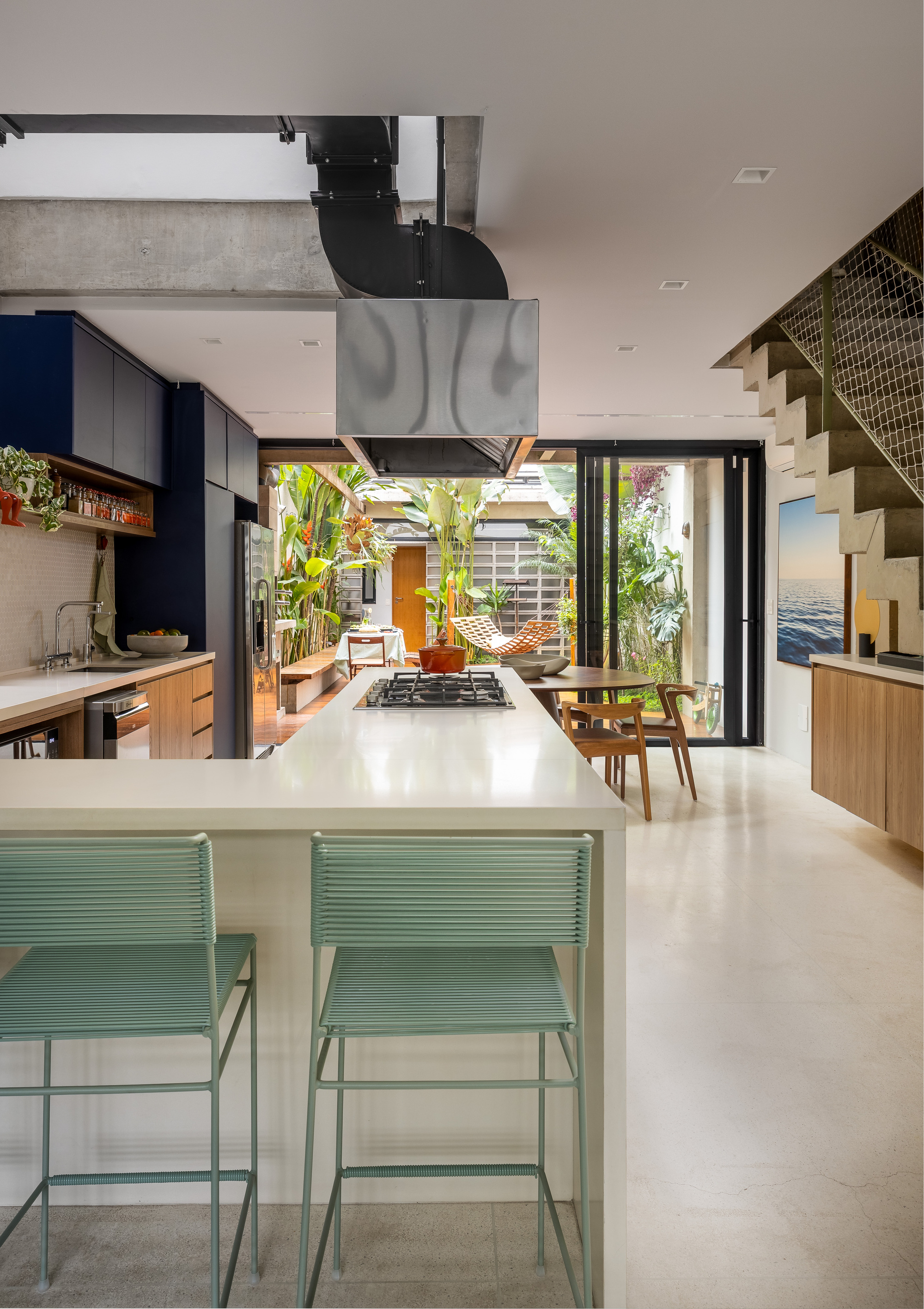 Kitchen island with a gas hob and two mint green stools, looking through open glass doors to a leafy courtyard