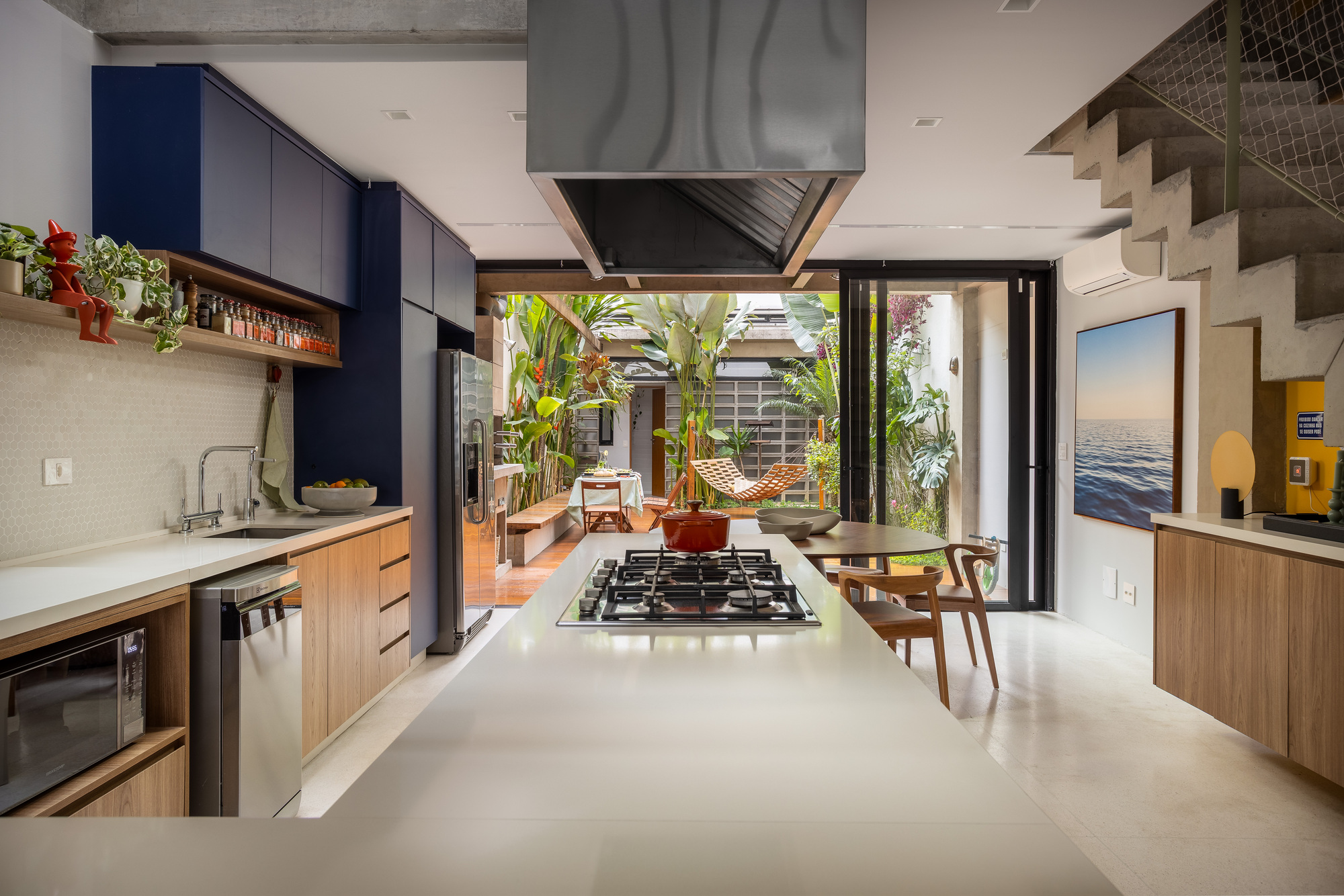 Long kitchen island with gas cooktop and red pot, opening through folding glass doors to a planted courtyard in daylight