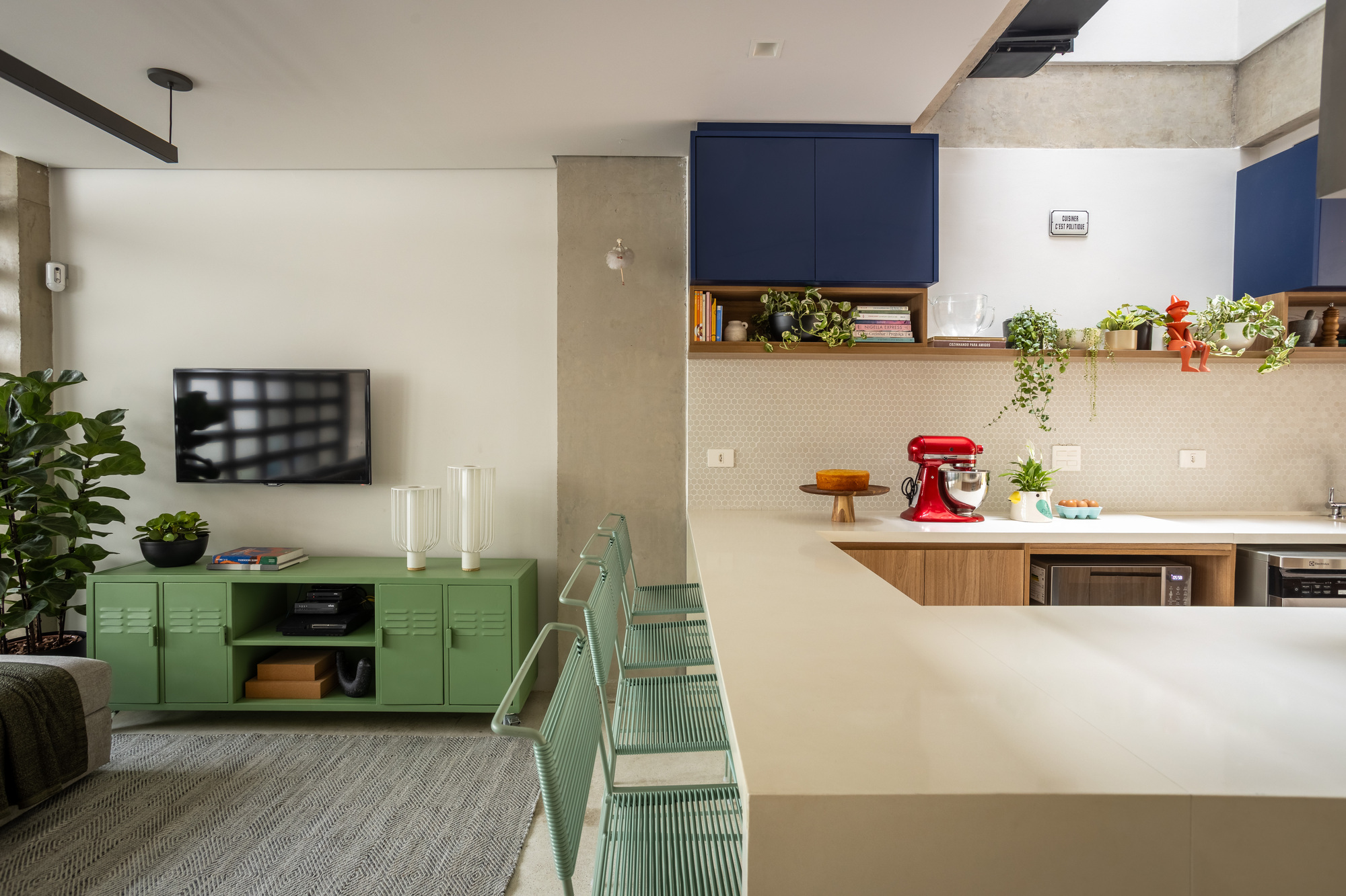 Kitchen peninsula with mint wire stools beside a green locker-style media unit and a concrete column in soft daylight
