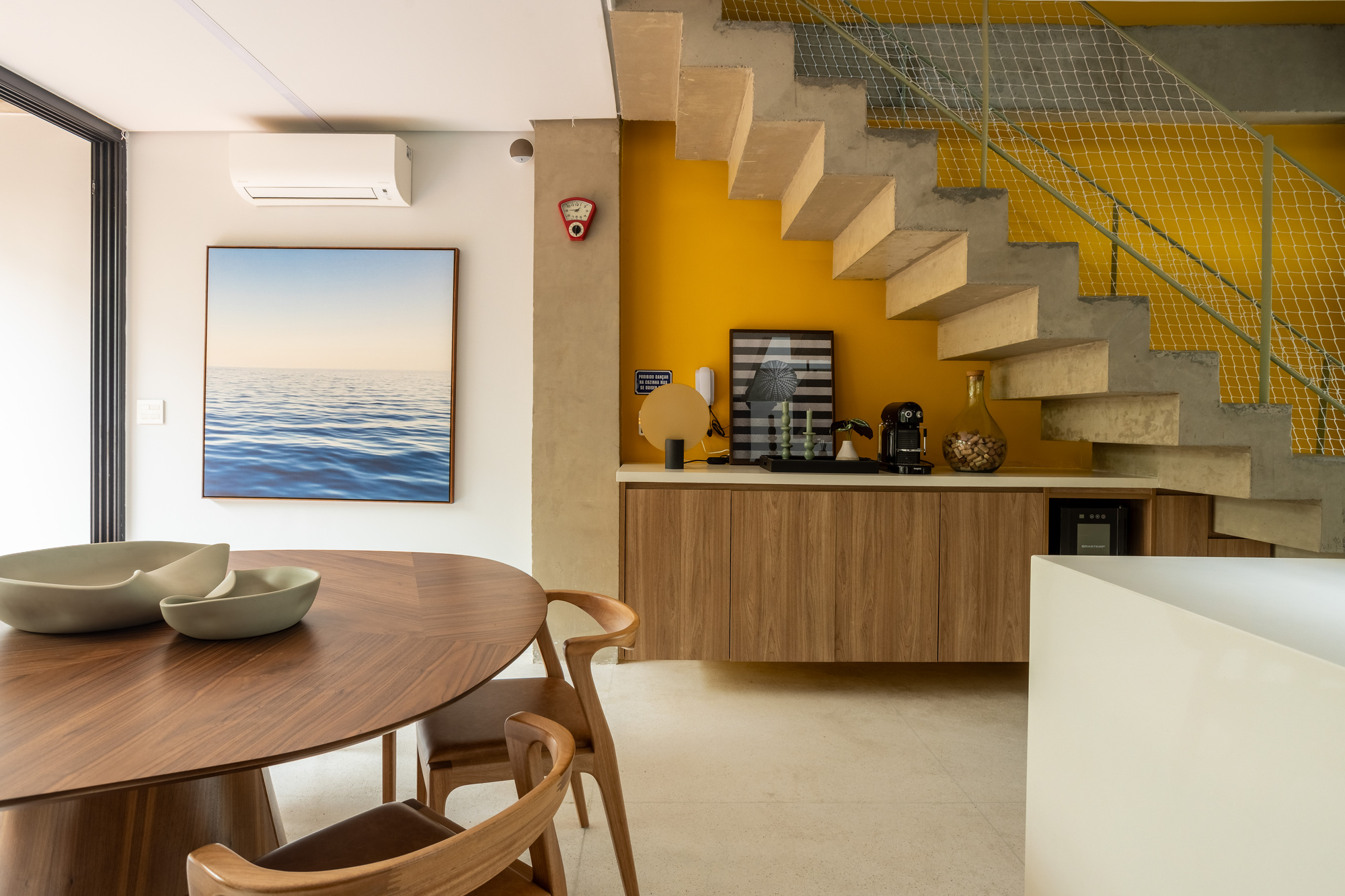 Concrete stair with cantilevered treads above a floating timber sideboard against a mustard yellow wall, beside a round walnut dining table