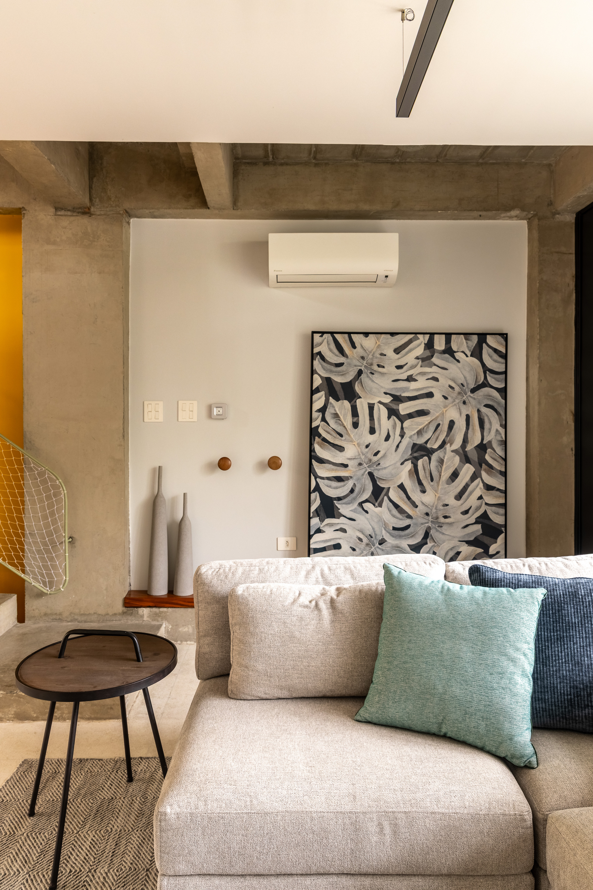 Living room corner with a gray sofa, teal and blue cushions and a monstera-leaf print against exposed concrete beams