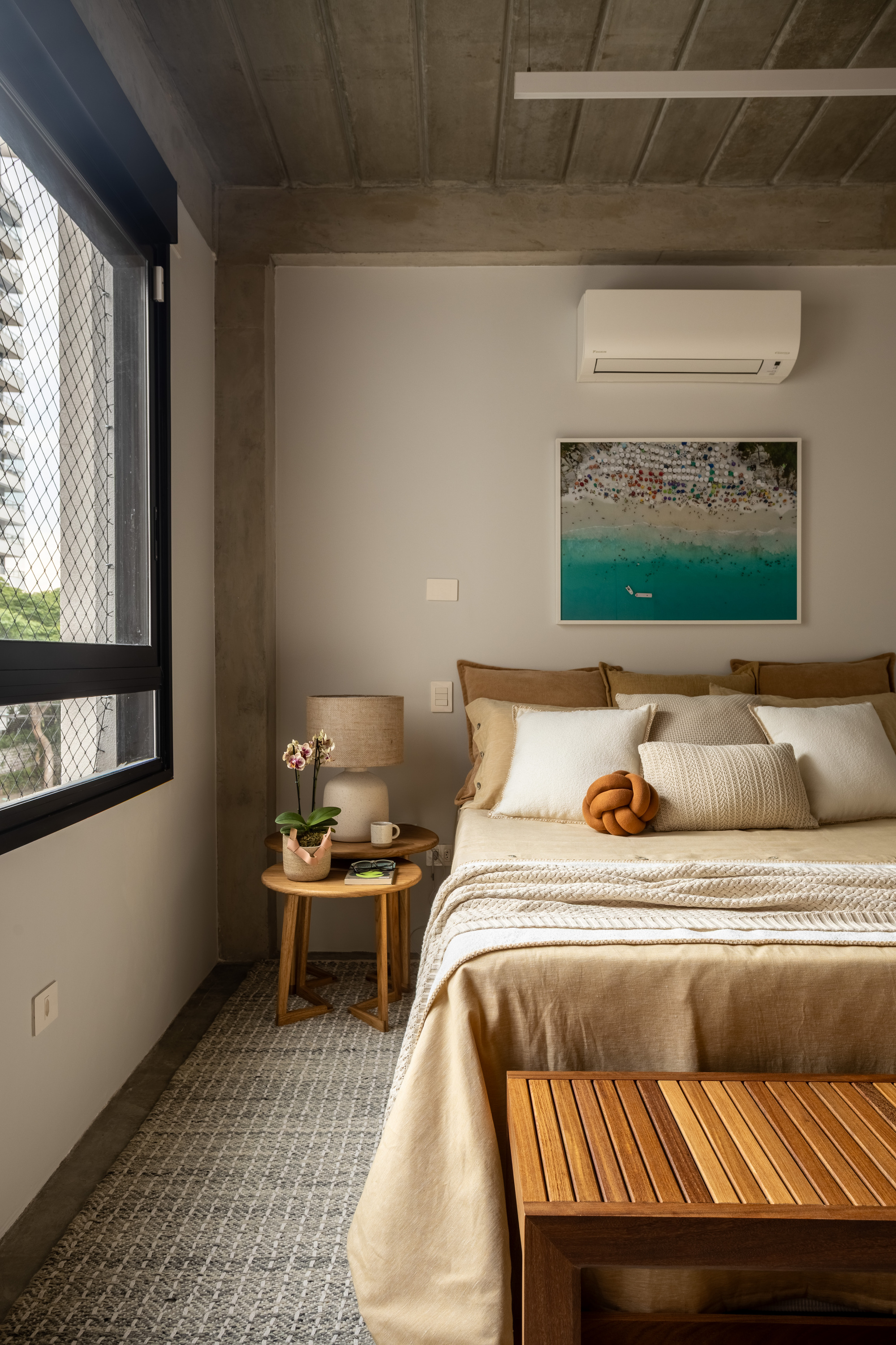 Bedroom with a linen-covered bed, a round timber side table and a window screened by steel mesh onto neighboring towers