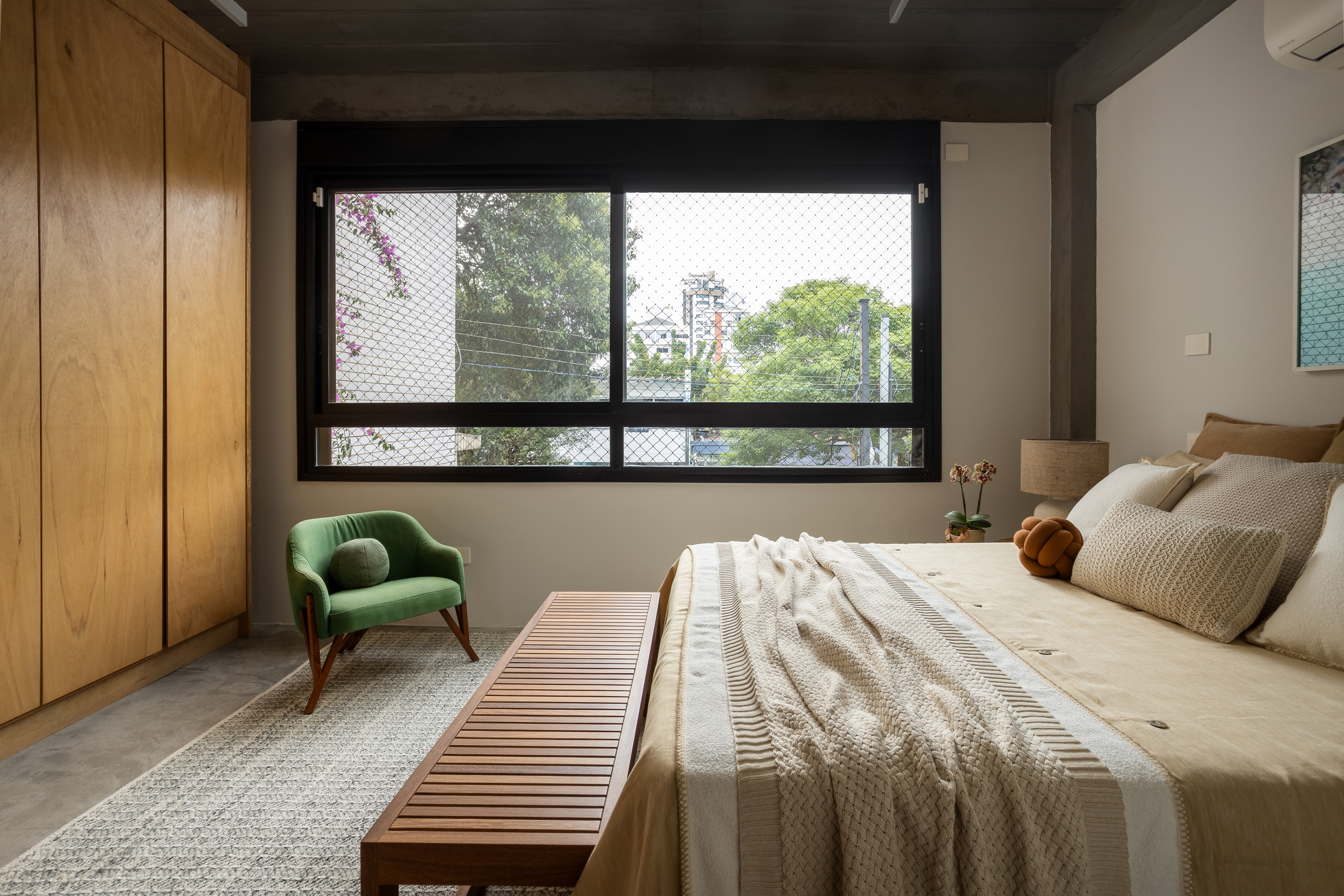 Bedroom seen across a slatted wooden bench, with a green armchair and a mesh-screened window framing tree canopy in daylight