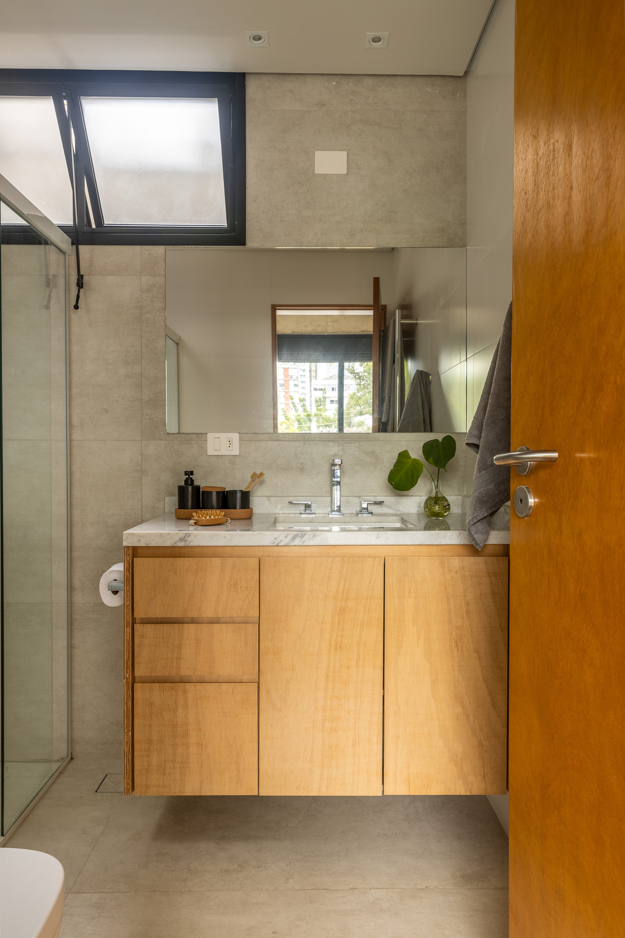 Bathroom with a floating oak vanity, marble countertop and a mirror reflecting a window, beside a glass shower screen