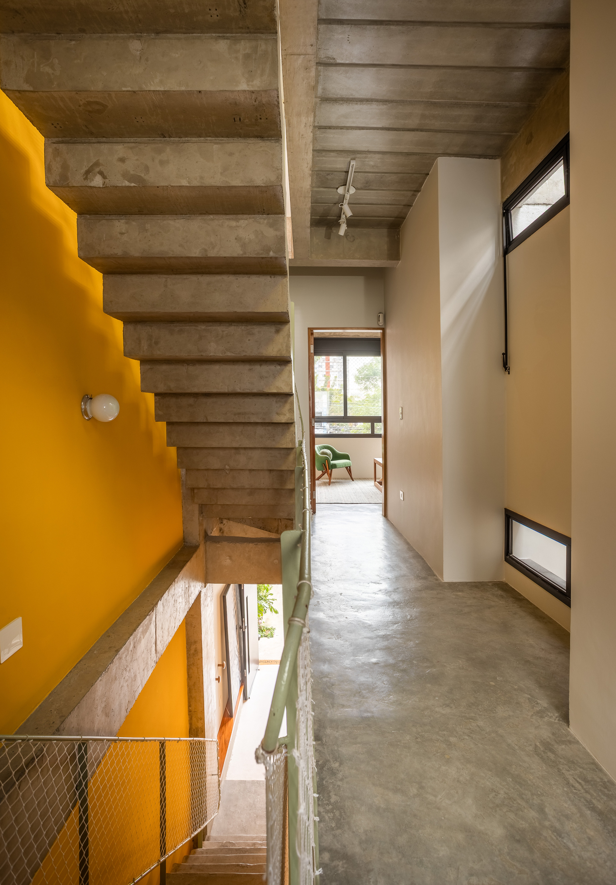 Exposed concrete stair against a mustard yellow wall, with a mesh guardrail and a corridor leading to a bedroom