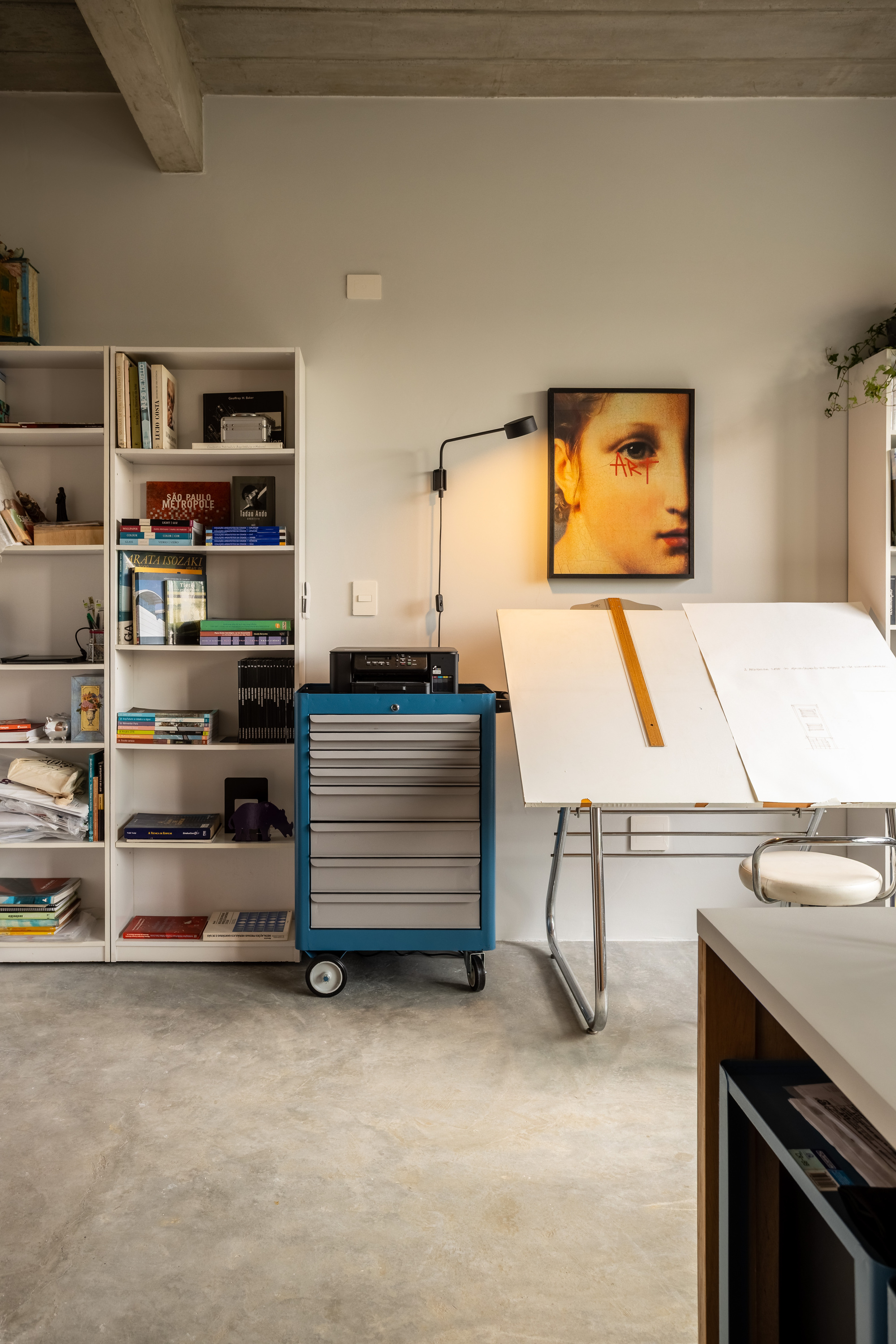 Studio corner with a drafting table, a blue rolling tool chest and white bookshelves under a concrete beam ceiling