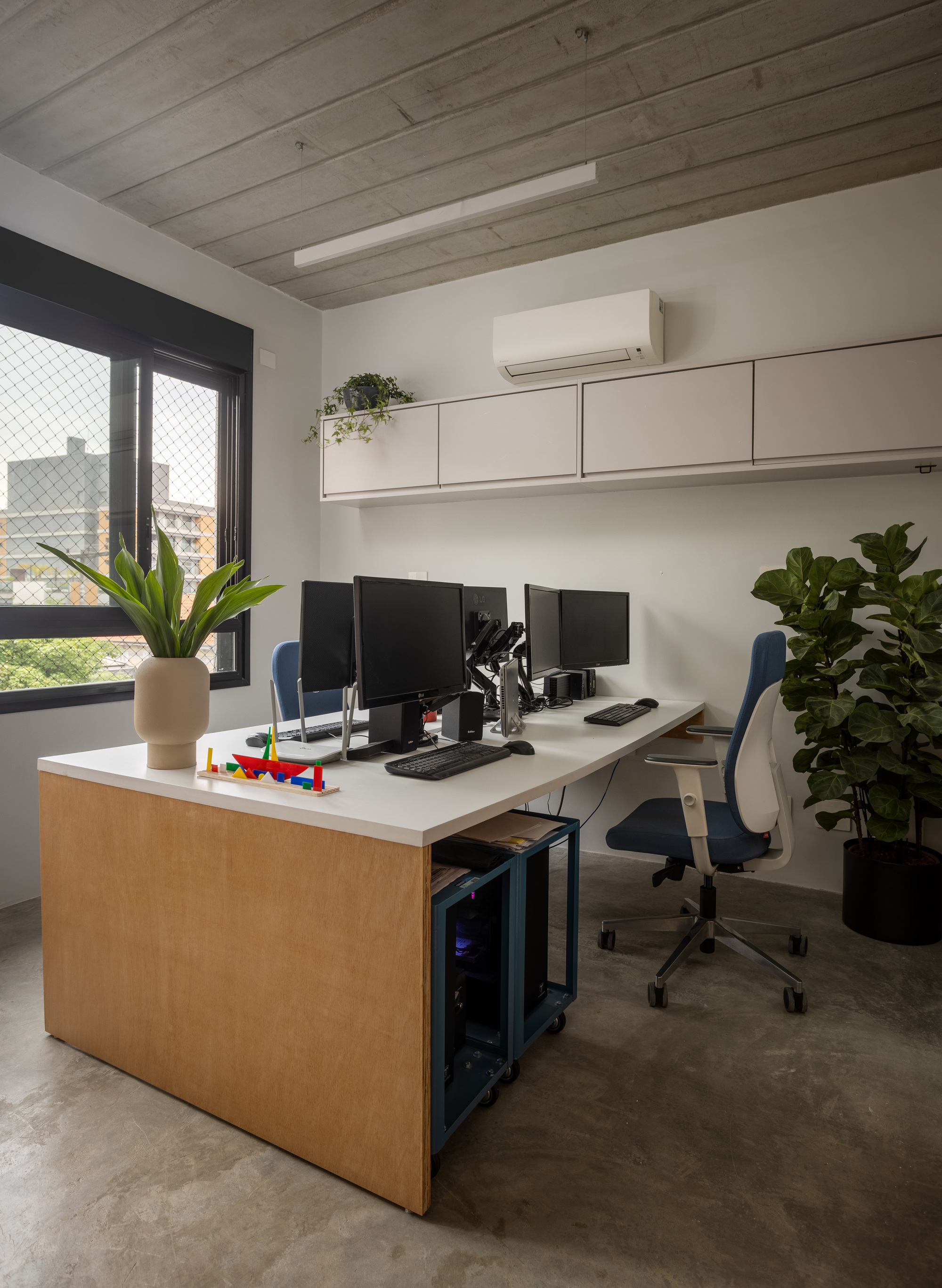 Office corner with a wood-sided white desk, dual monitors and potted plants beside a window, under a board-marked concrete ceiling