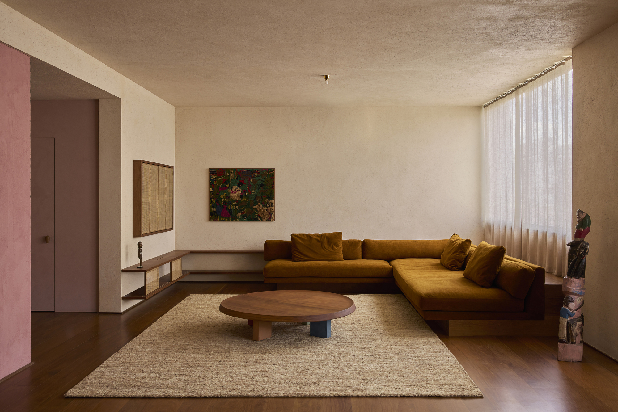 Living room with a mustard L-shaped sofa, round wood coffee table and jute rug beside sheer curtains in soft daylight