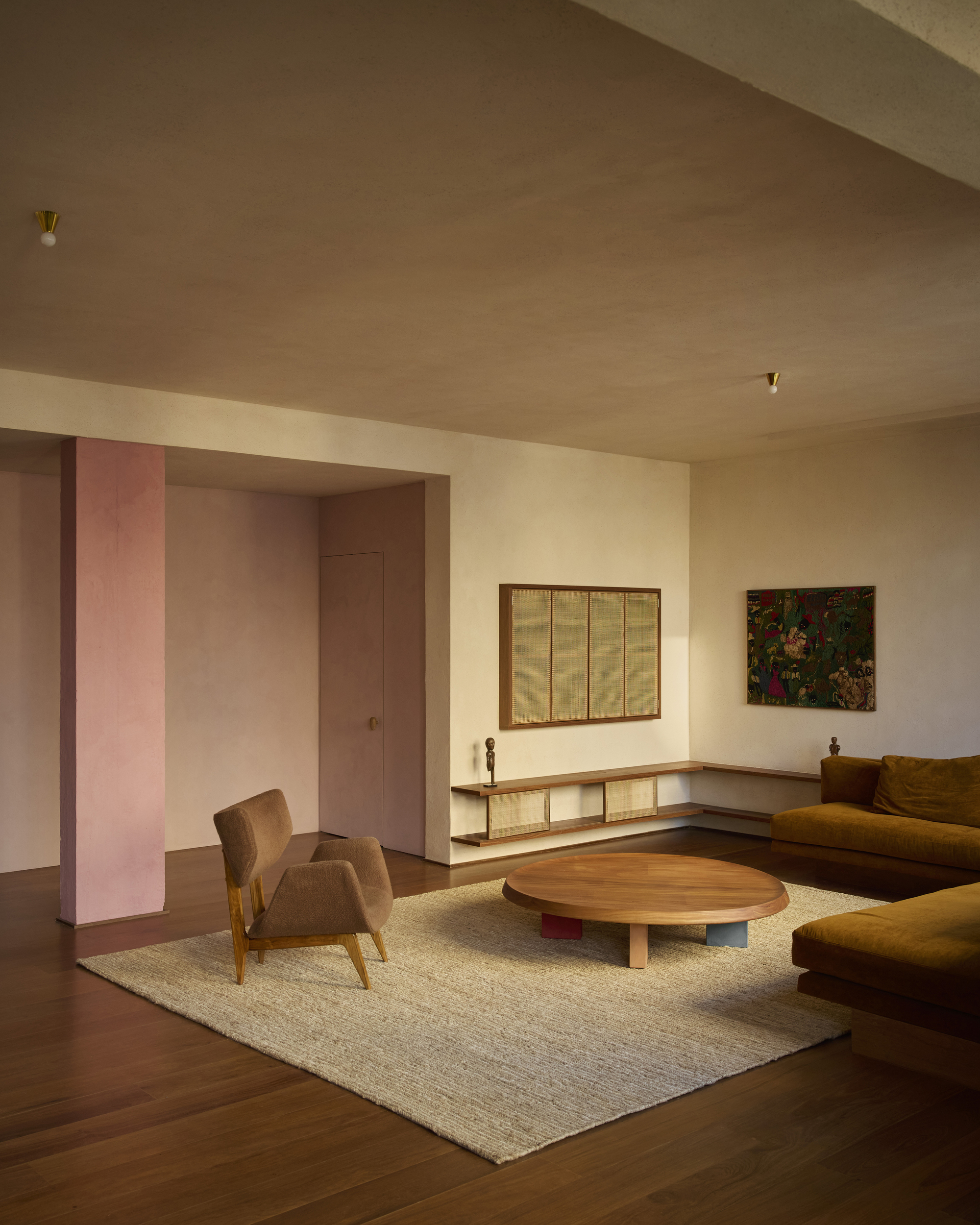 Lounge corner with a pink plastered column, cane-panelled wall cabinet and low timber shelf in warm late light