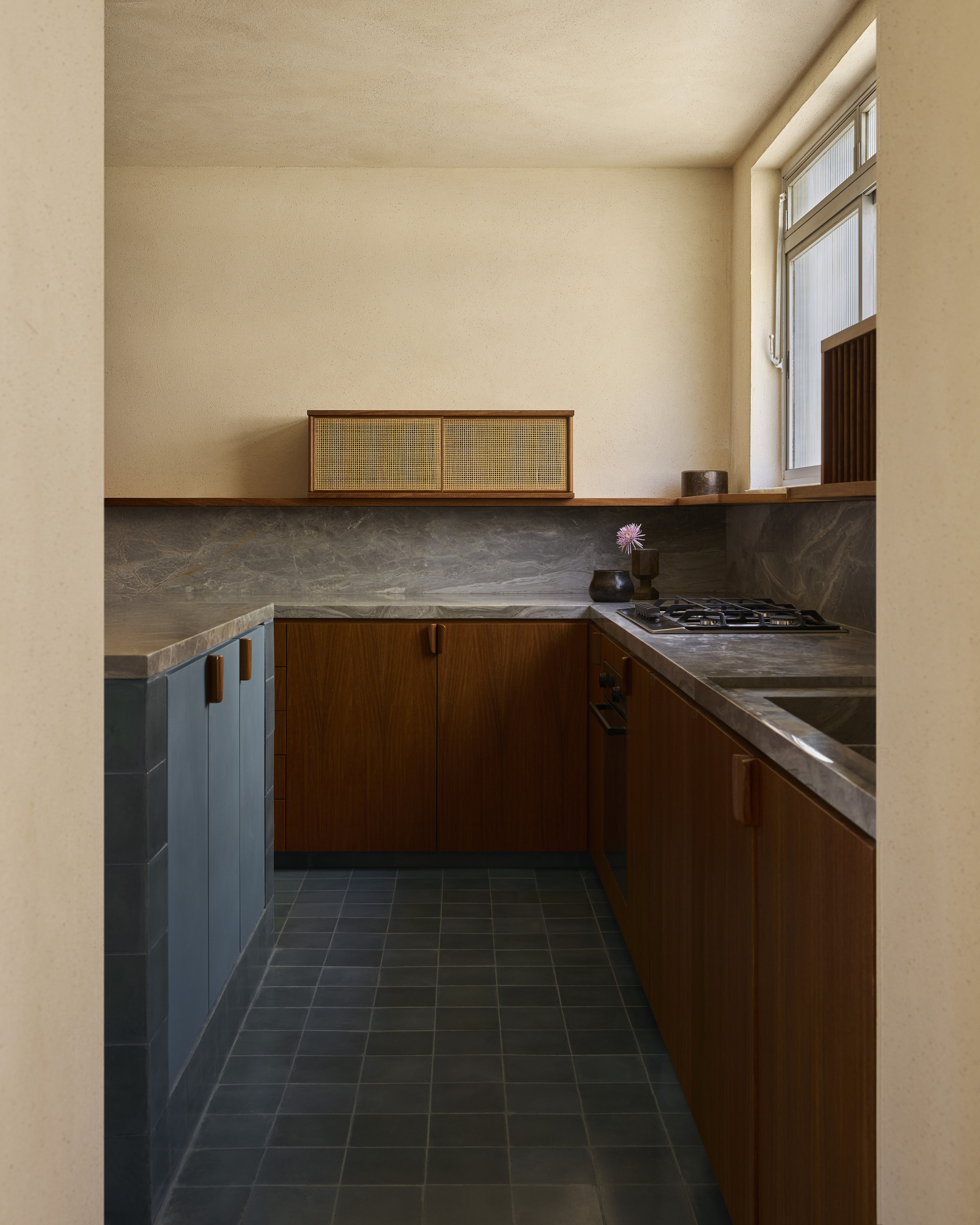 Galley kitchen with walnut cabinets, grey stone backsplash and blue-grey tiled base units under daylight from a side window