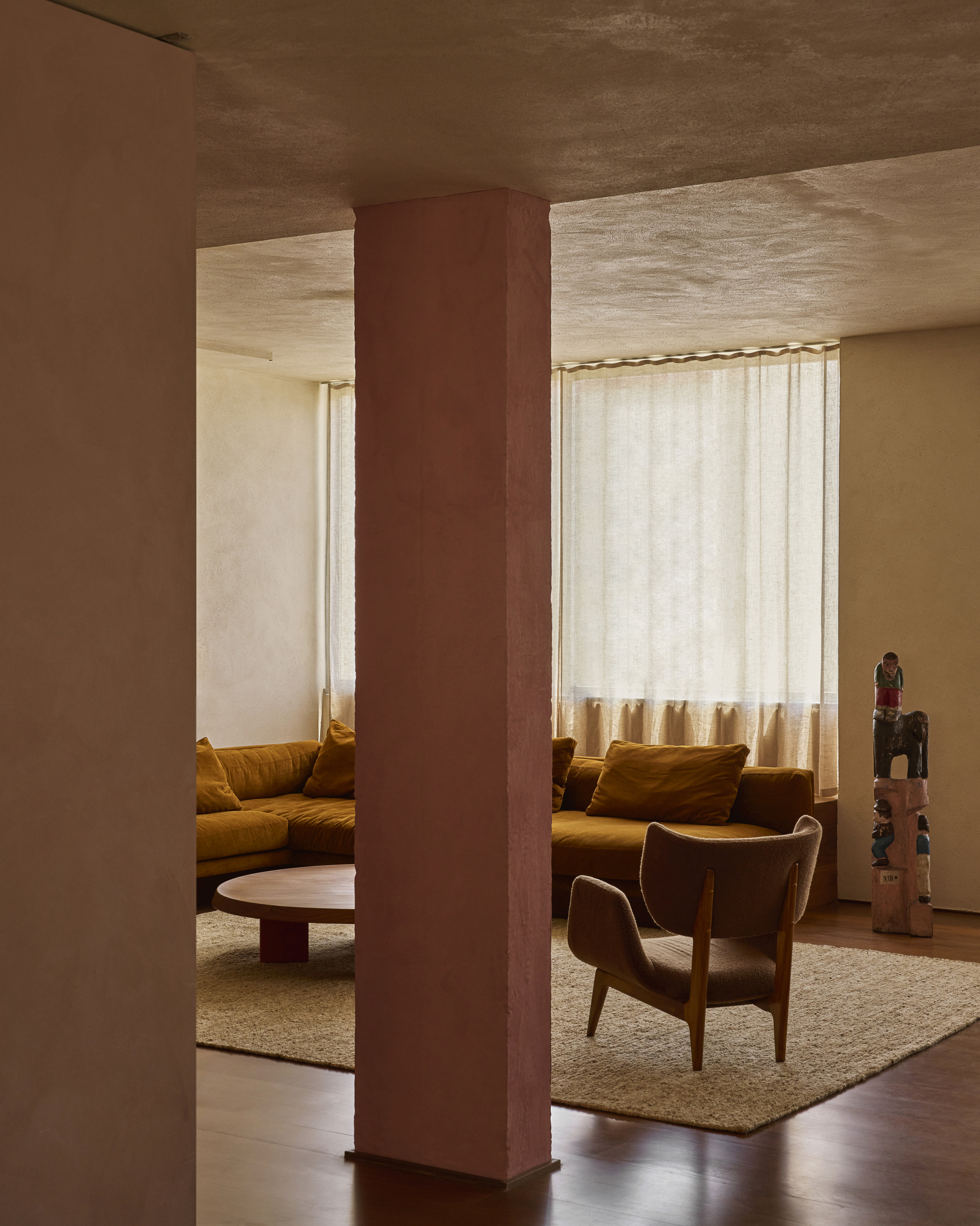 Pink plastered column framing the mustard sofa, wood armchair and carved totem figure behind sheer curtains in afternoon light