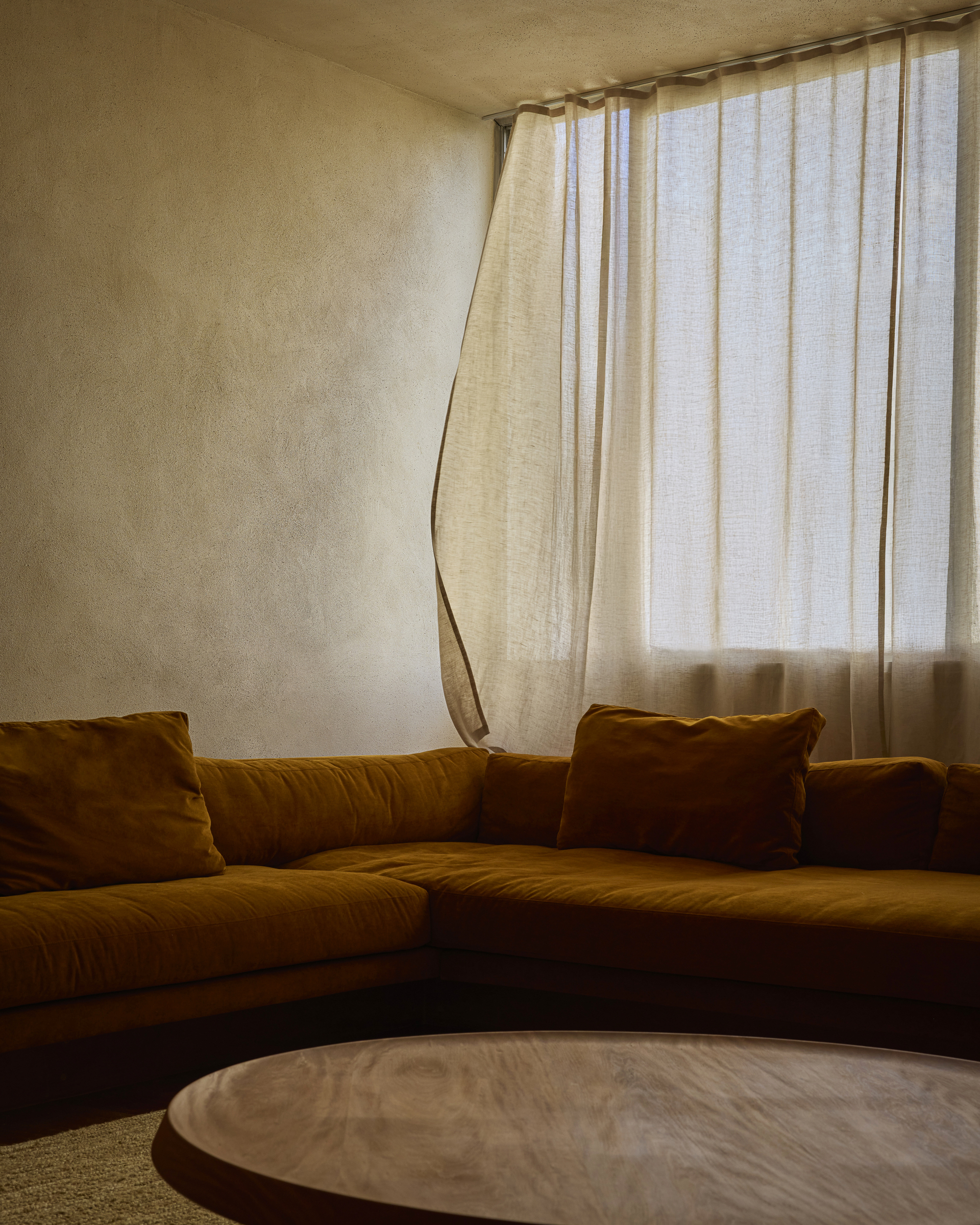 Close-up of the mustard velvet sofa corner and a linen curtain stirred by breeze beside a round timber table