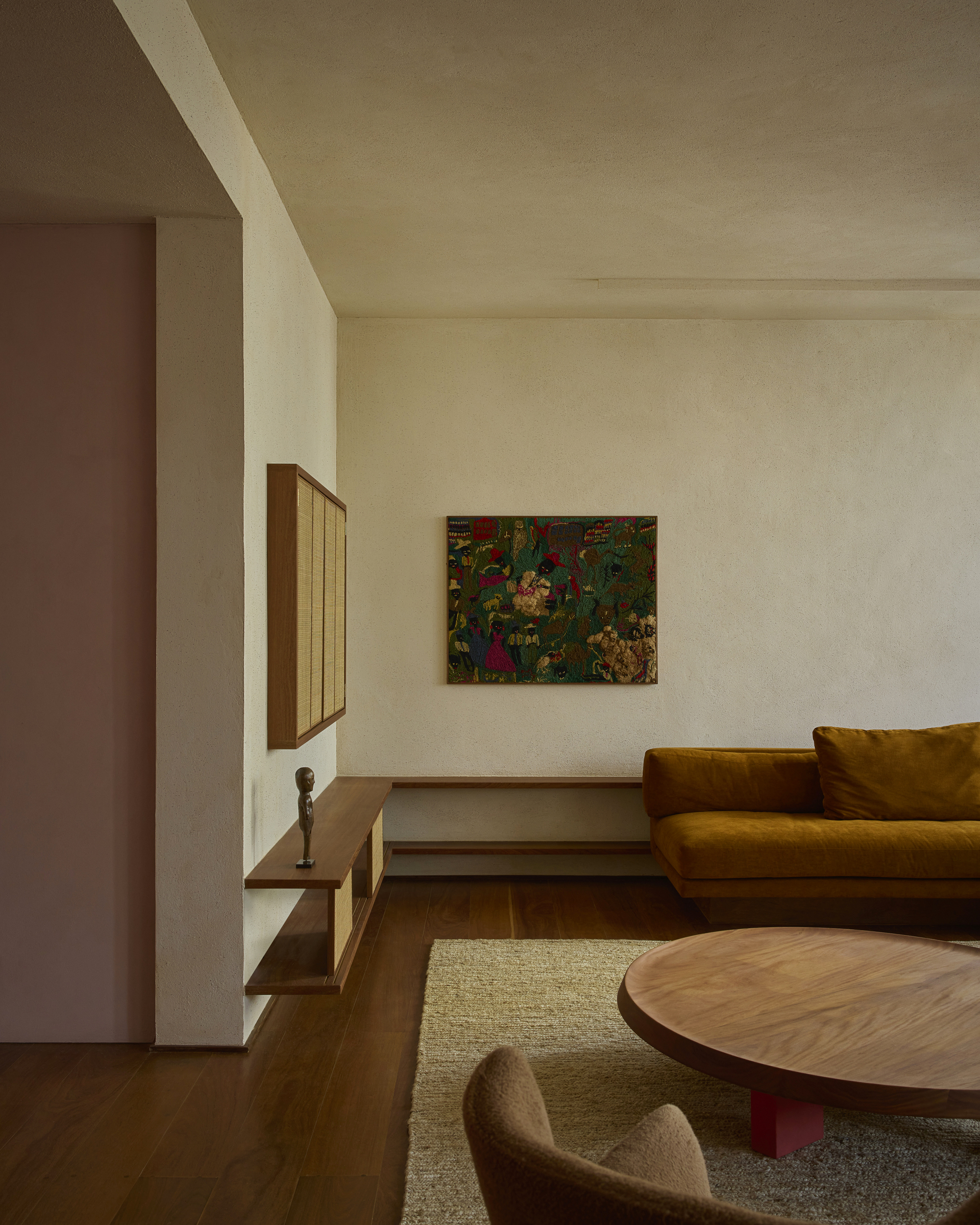 Living room corner with a mustard velvet sofa, round walnut coffee table and a framed textile on a plastered wall in low light