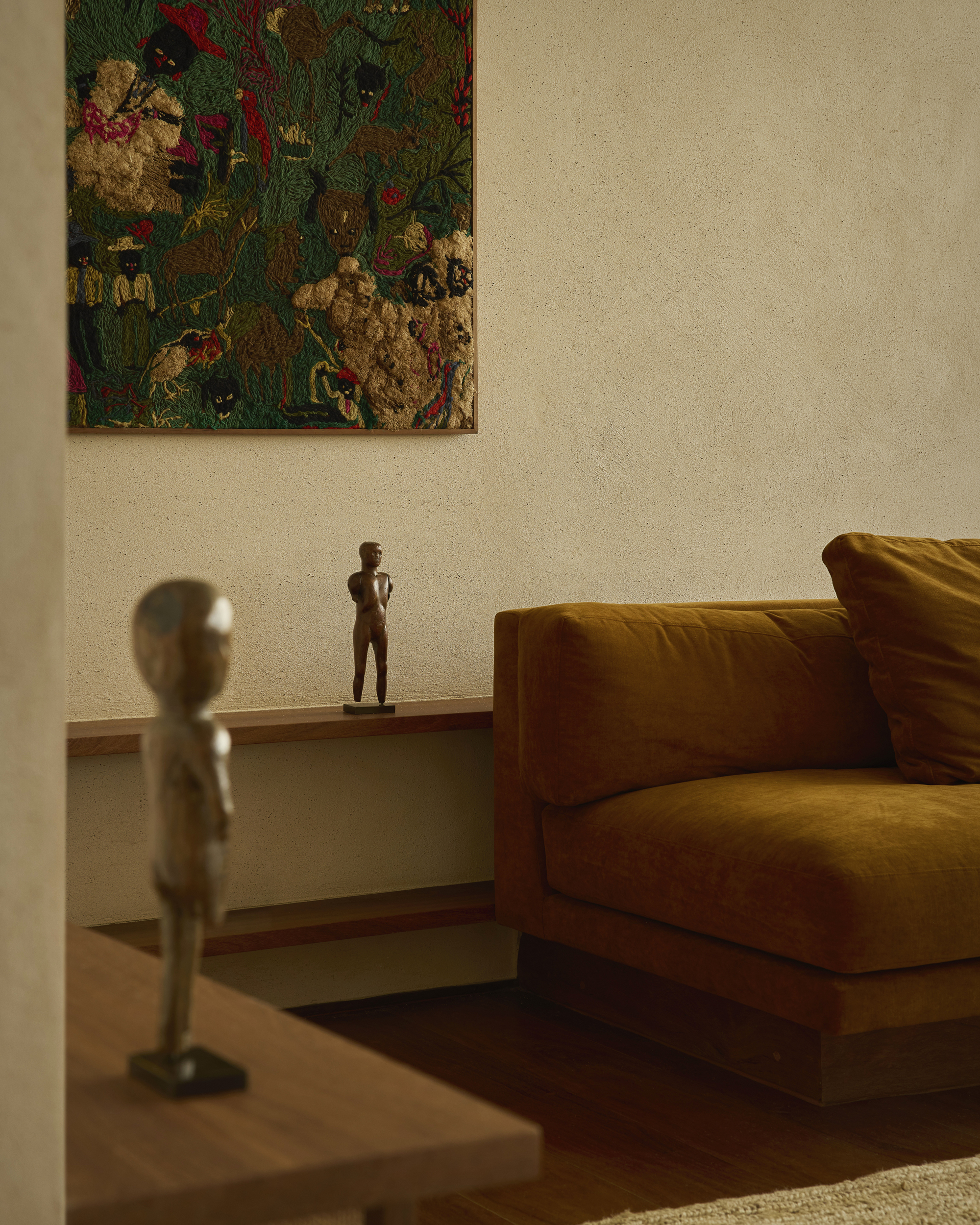 Close view of a small bronze figure on a wall shelf beside a velvet sofa, with a woven textile above in warm light