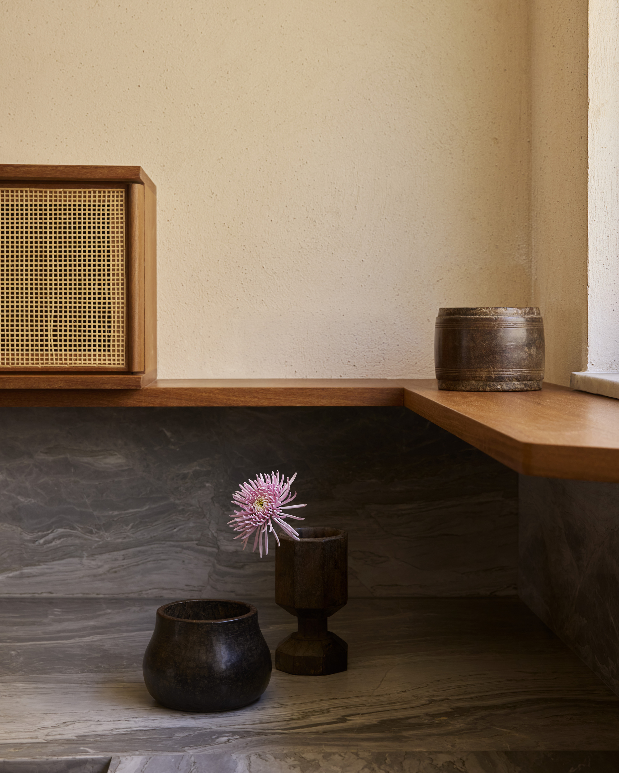 Corner of a timber shelf above a grey marble ledge holding dark wooden vessels and a pink chrysanthemum, with window light at right