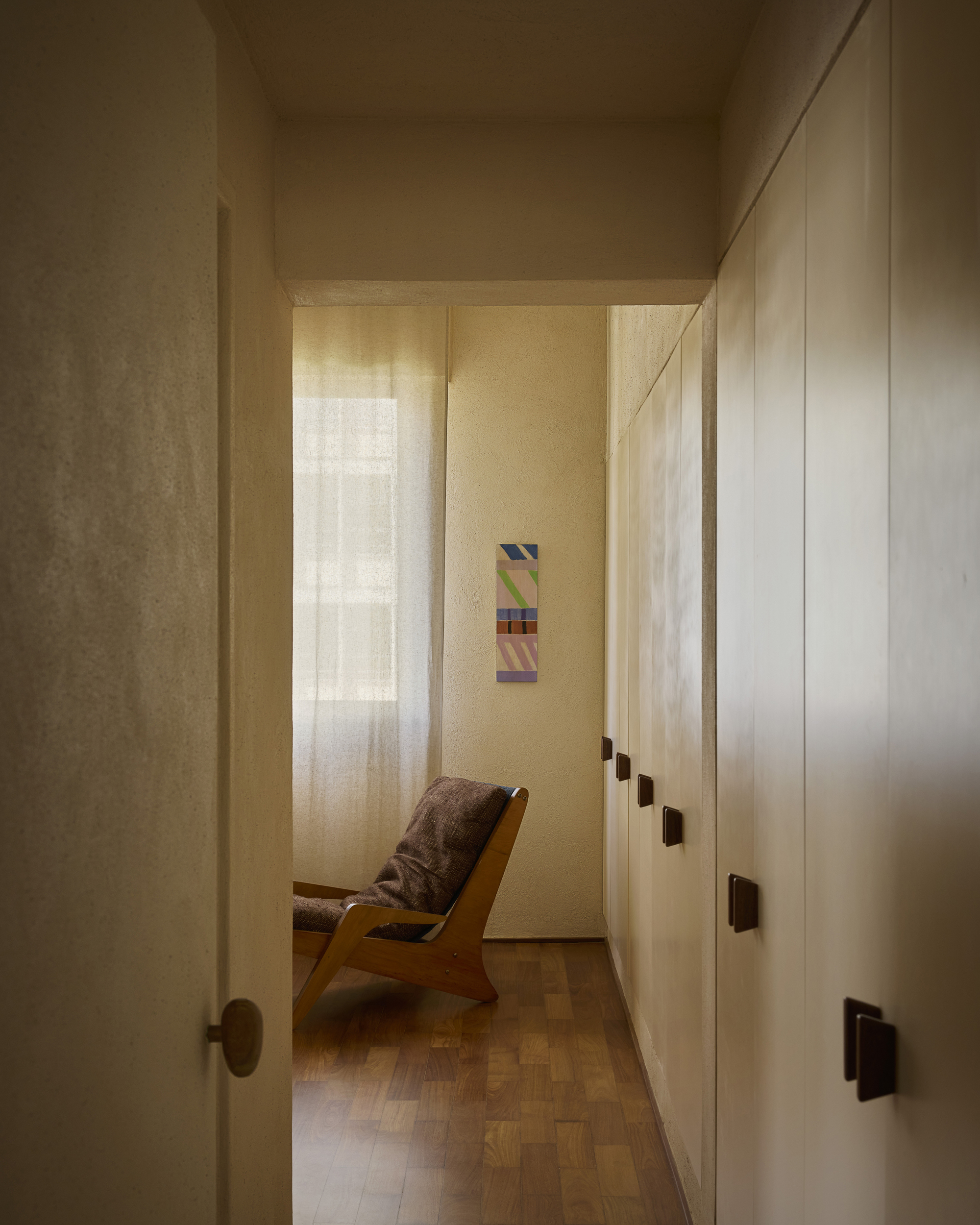 Narrow hallway lined with white wardrobe doors and wooden handles, leading to a lounge chair in front of a sheer curtain