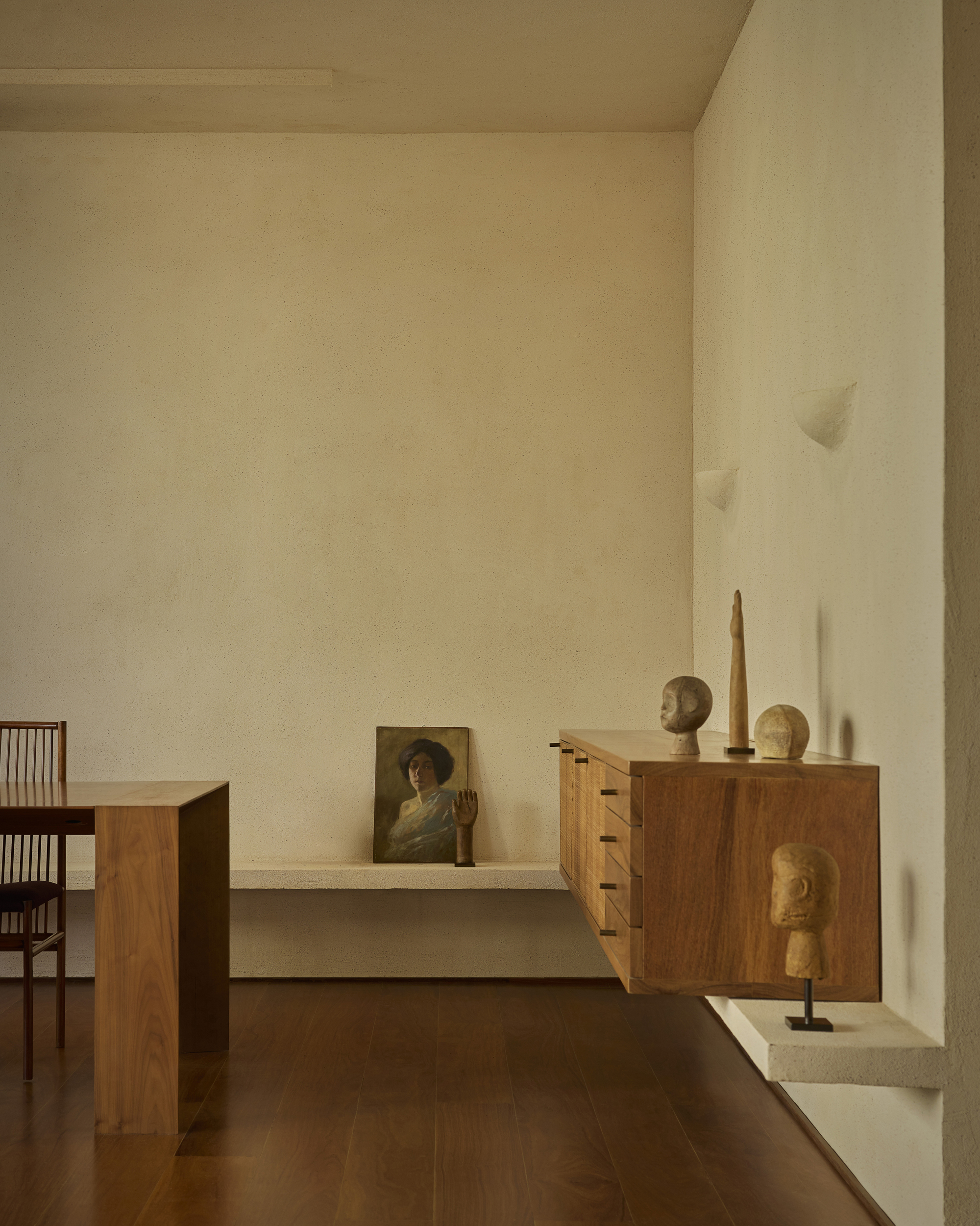 Floating timber sideboard topped with carved heads and a figure, beside a dining table and a framed portrait resting on a plaster ledge