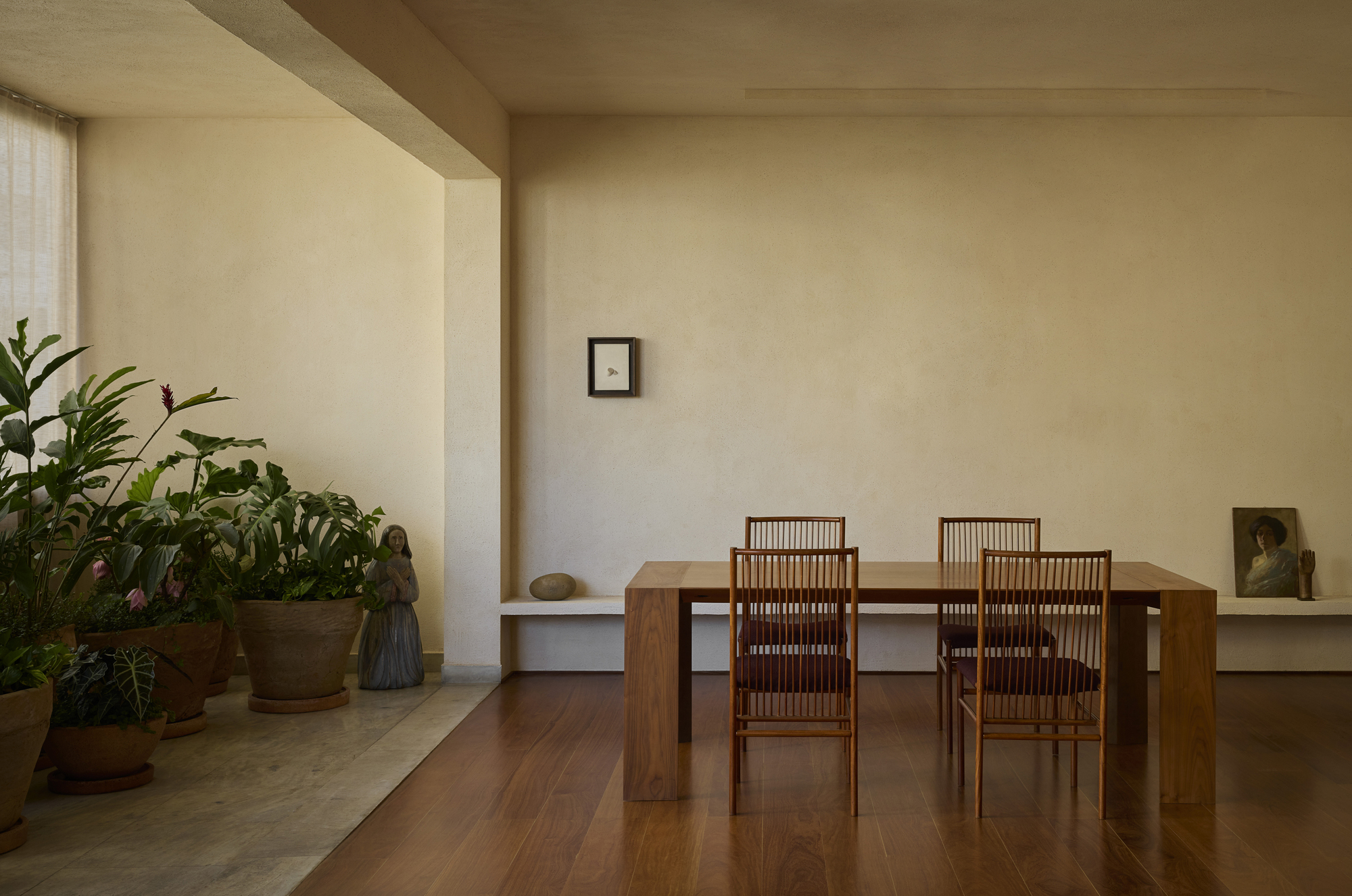 Dining room with a timber table and four slatted chairs, beside a stone-floored alcove of potted tropical plants in soft daylight