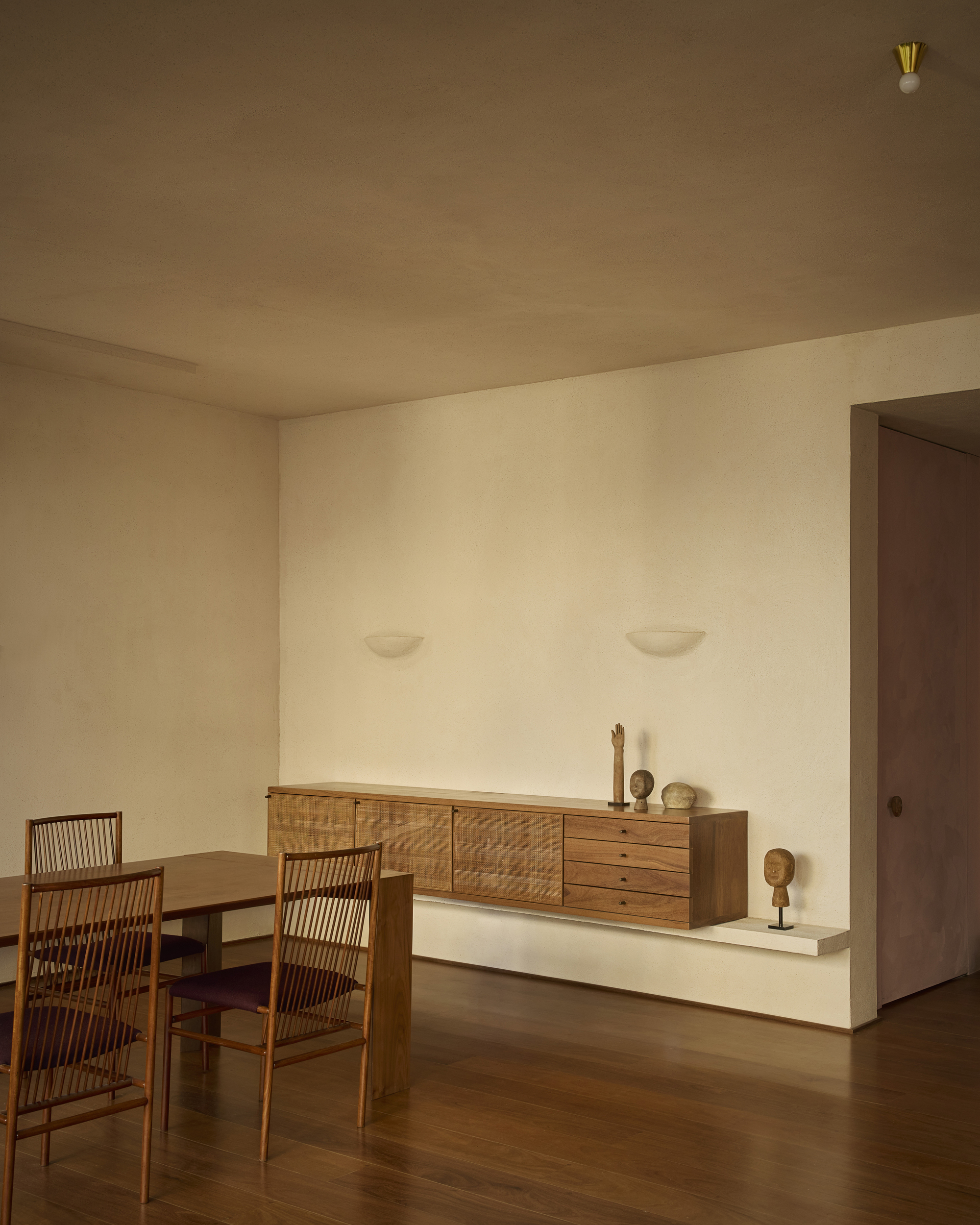 Floating timber sideboard with small carved heads and stone objects against a plastered wall, beside a dining table in low warm light