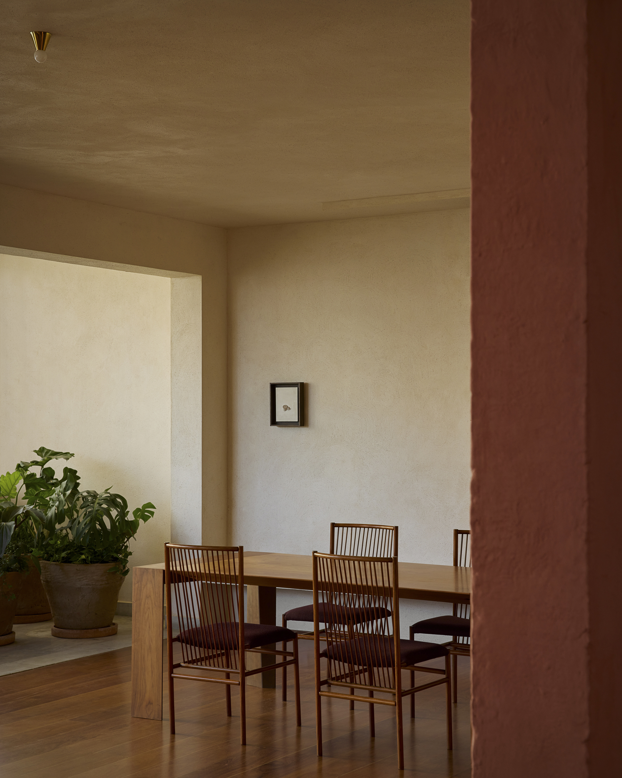 Dining table with spindle-back chairs seen past a pink plastered column, with potted plants in a side alcove in afternoon light