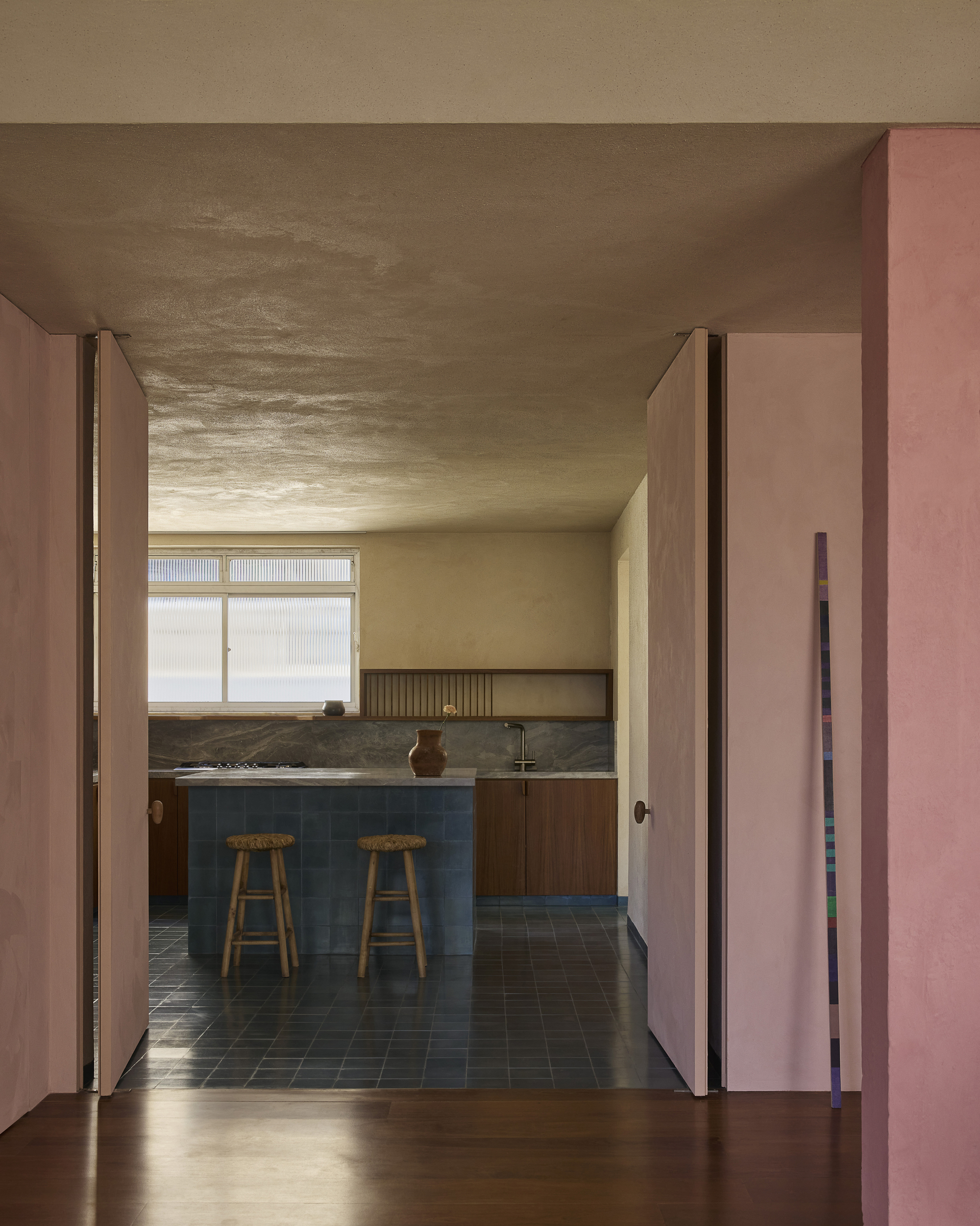 View through open pink pivot doors to a kitchen with blue tile island, two wooden stools and a ribbed window