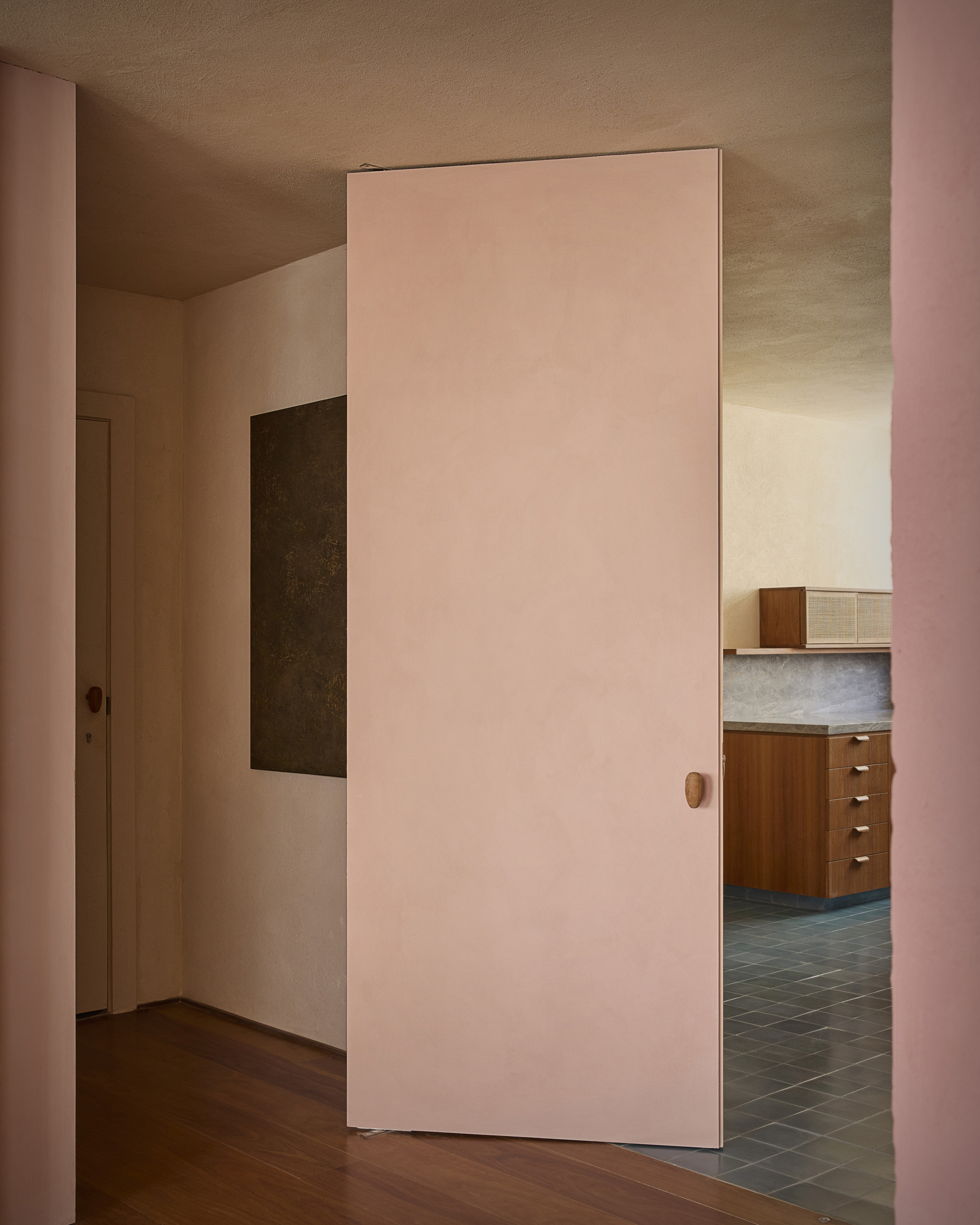 Pink pivot door with an oval wooden handle, standing open to reveal a walnut kitchen cabinet and blue-grey floor tiles
