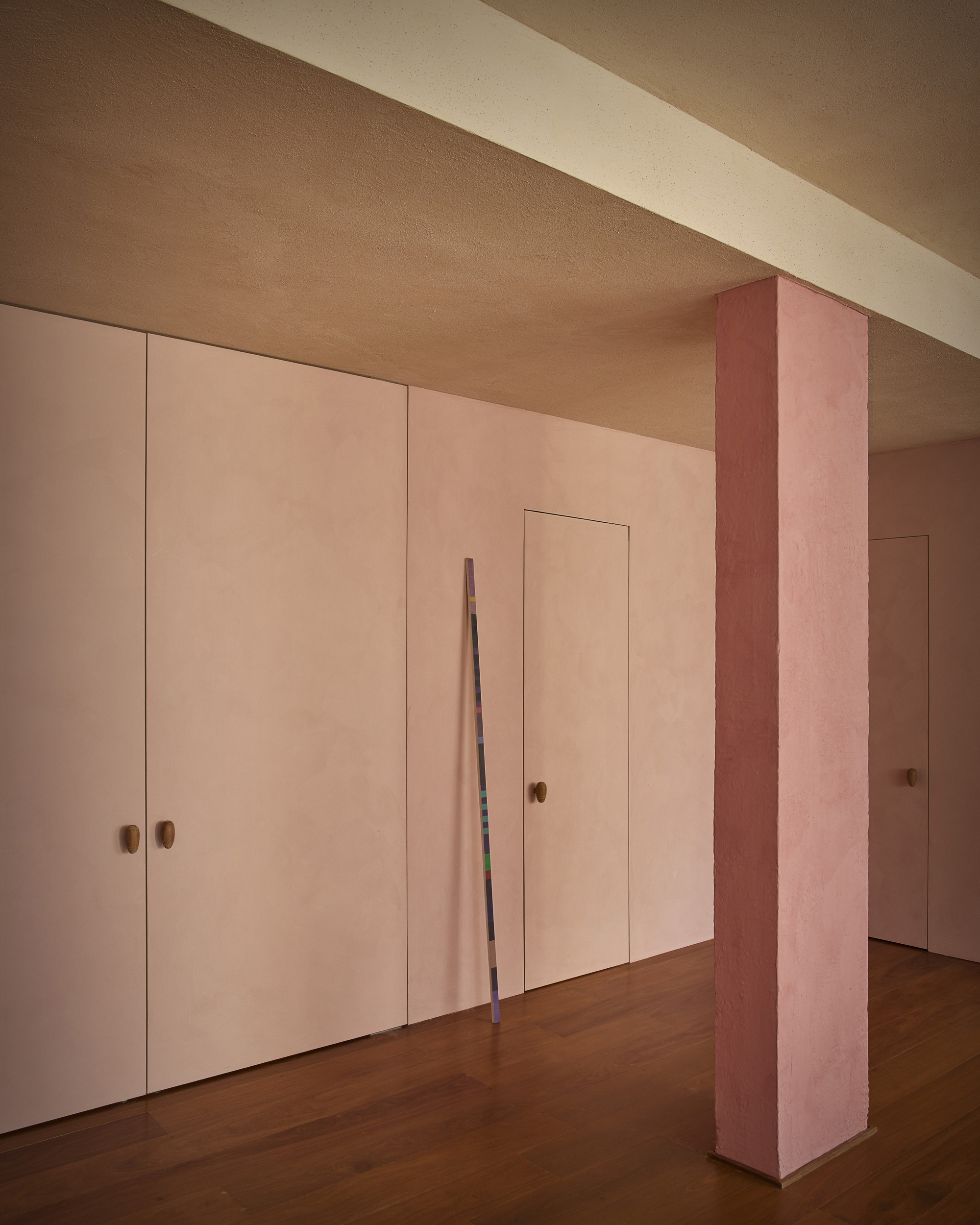 Flush pink doors with round wooden pulls beside a pink plastered column, a striped pole leaning on the wall in soft daylight