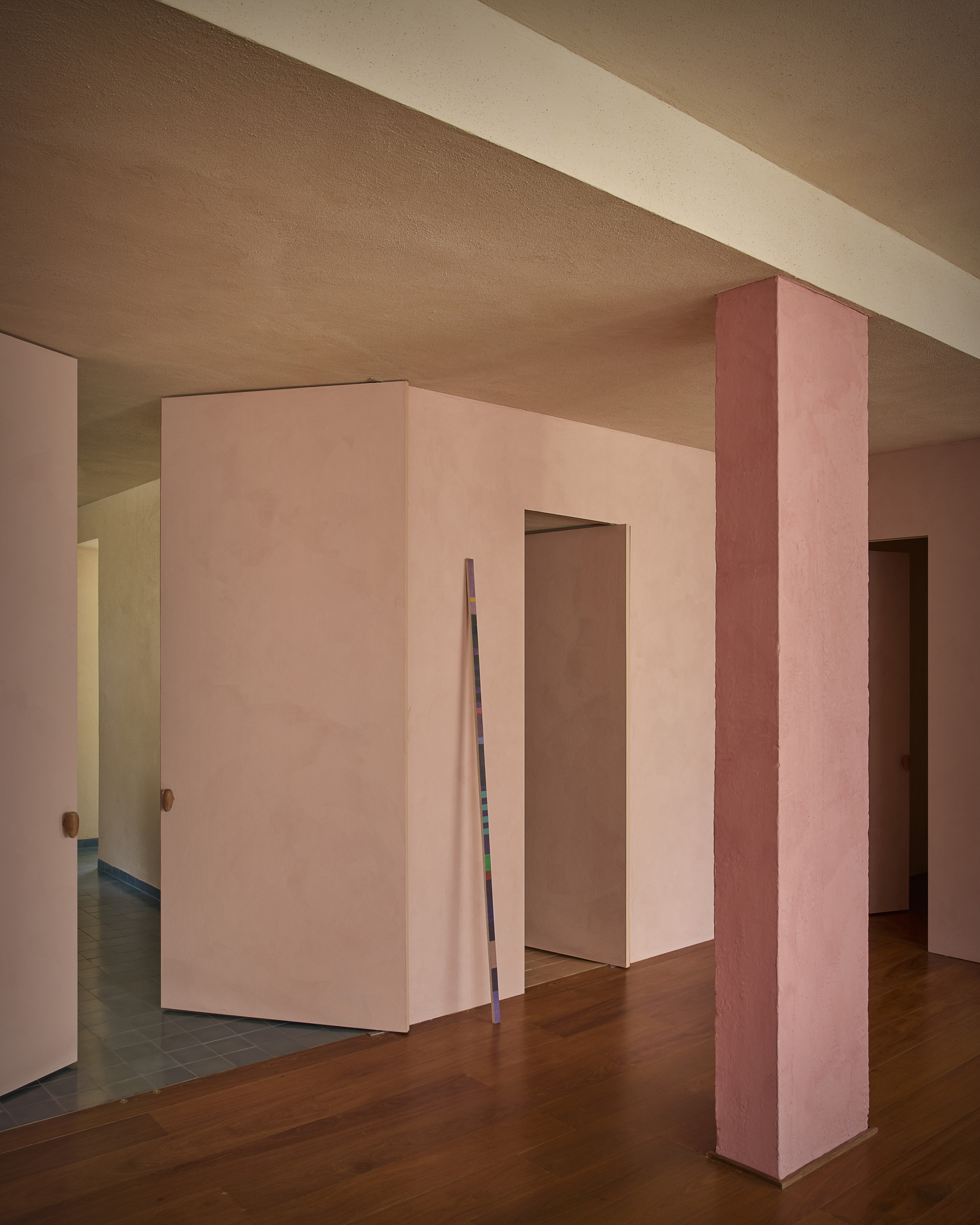 Pink plastered partition walls and a rough pink column over a timber floor, with a striped pole leaning in soft daylight