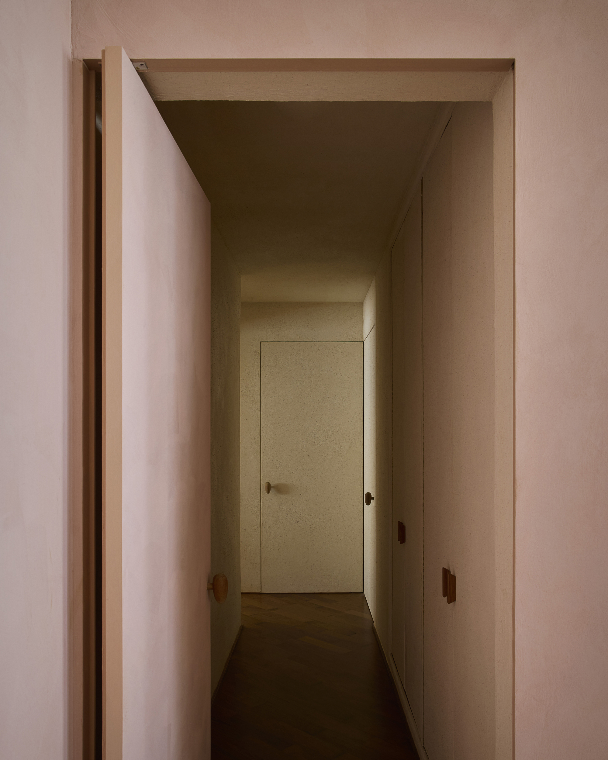 Narrow hallway seen through an open pink pivot door, with a flush white door at the end and built-in wardrobes