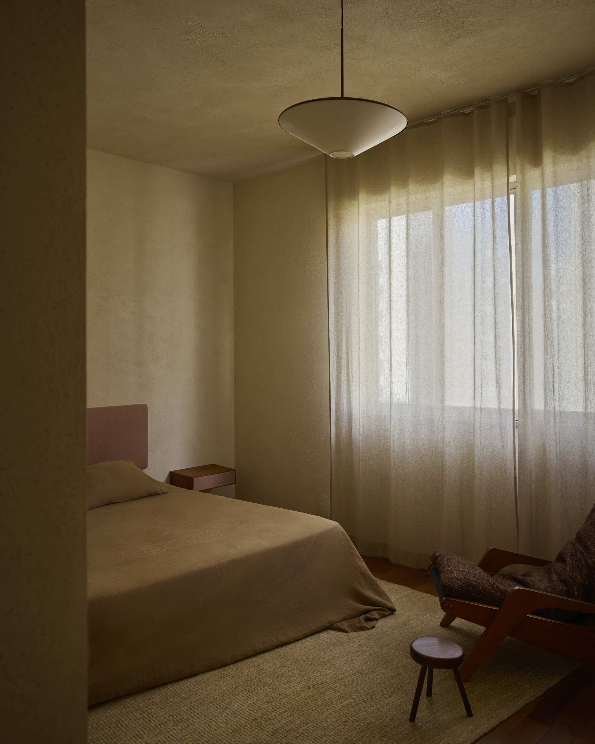 Bedroom with a low bed, conical pendant lamp and wooden lounge chair beside a sheer curtain filtering afternoon light