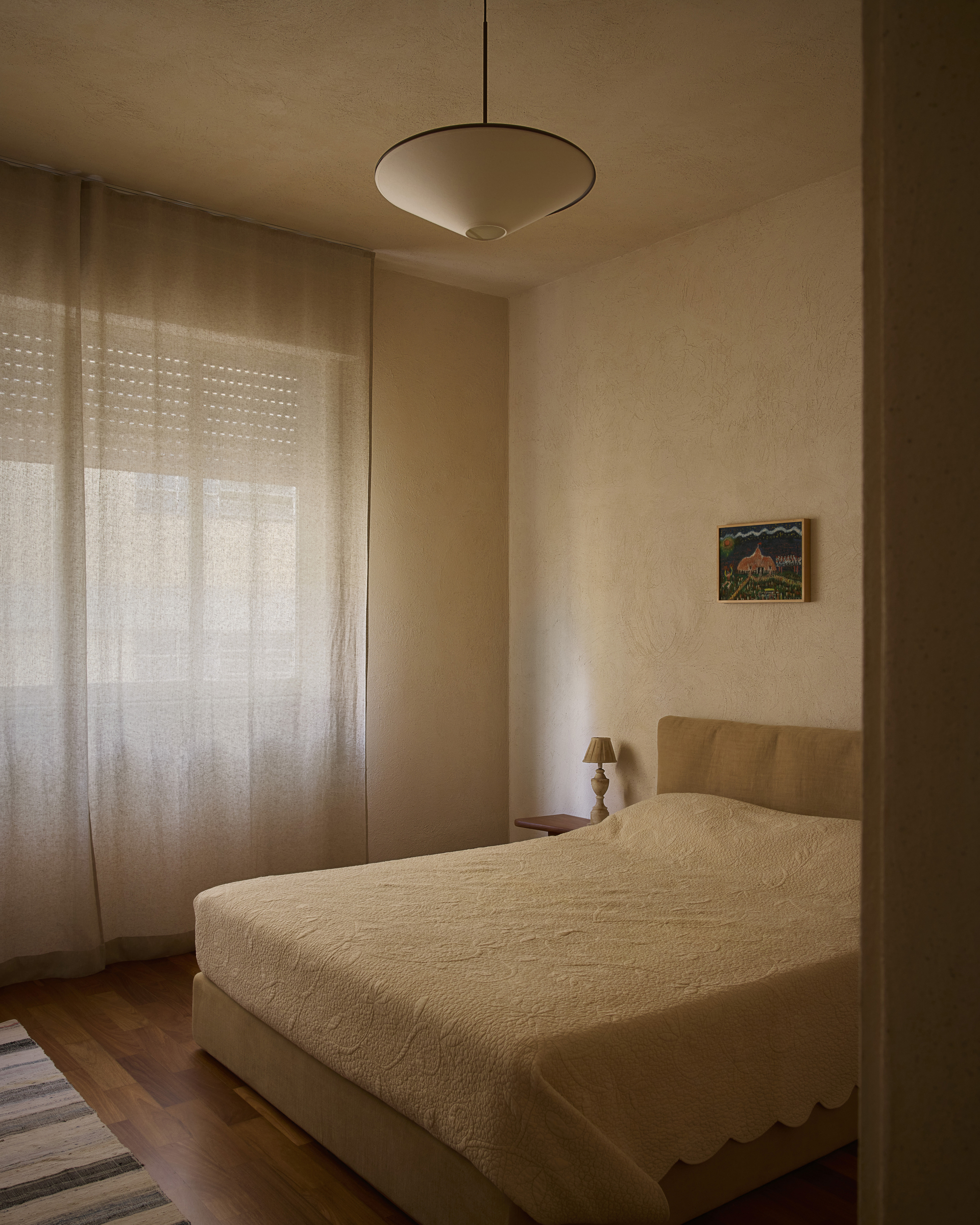 Bedroom with a quilted scalloped bedspread, small framed painting and bedside lamp beside a sheer-curtained window with roller shutter