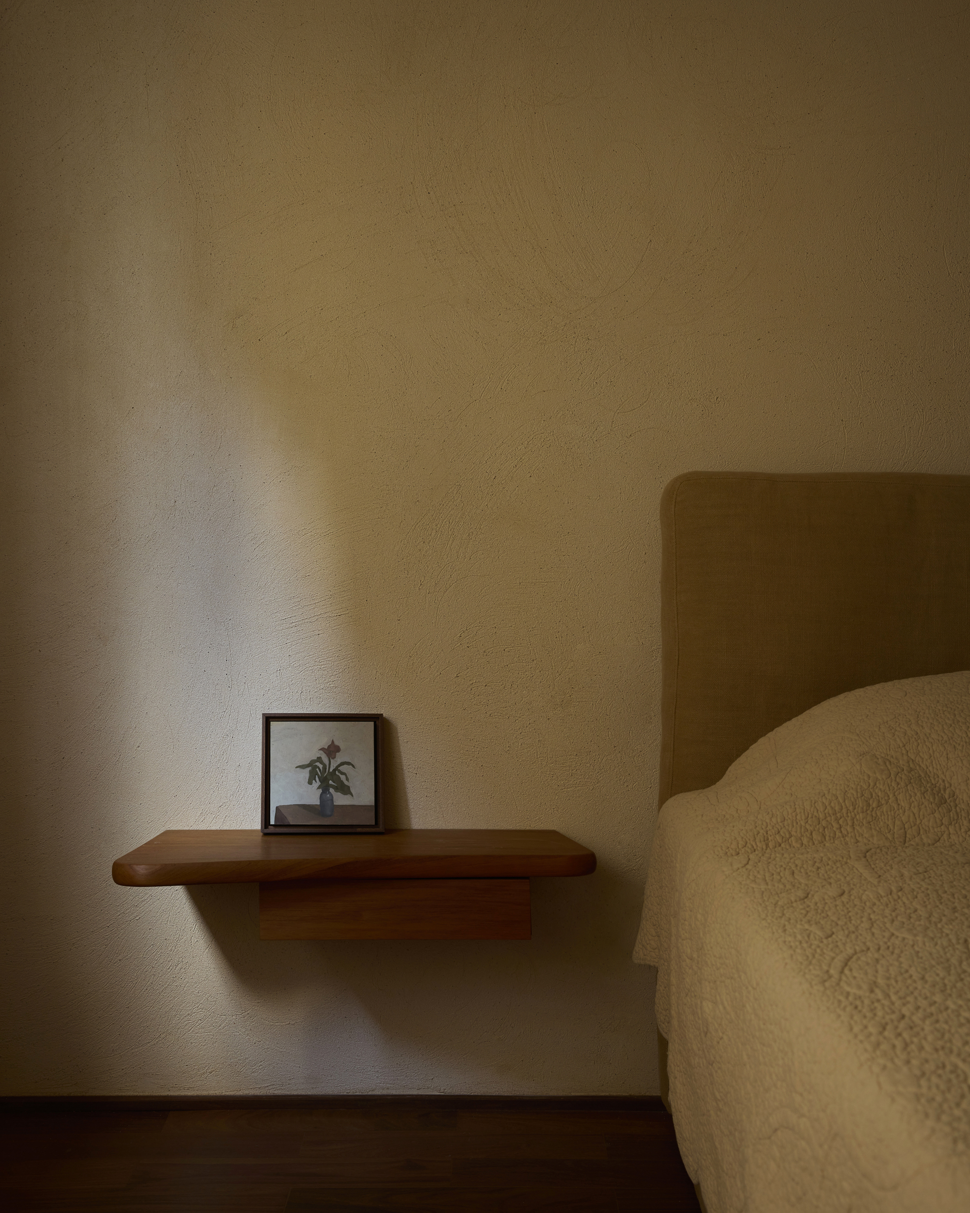 Wall-mounted timber bedside shelf holding a small framed flower painting, with a sliver of window light across the plaster
