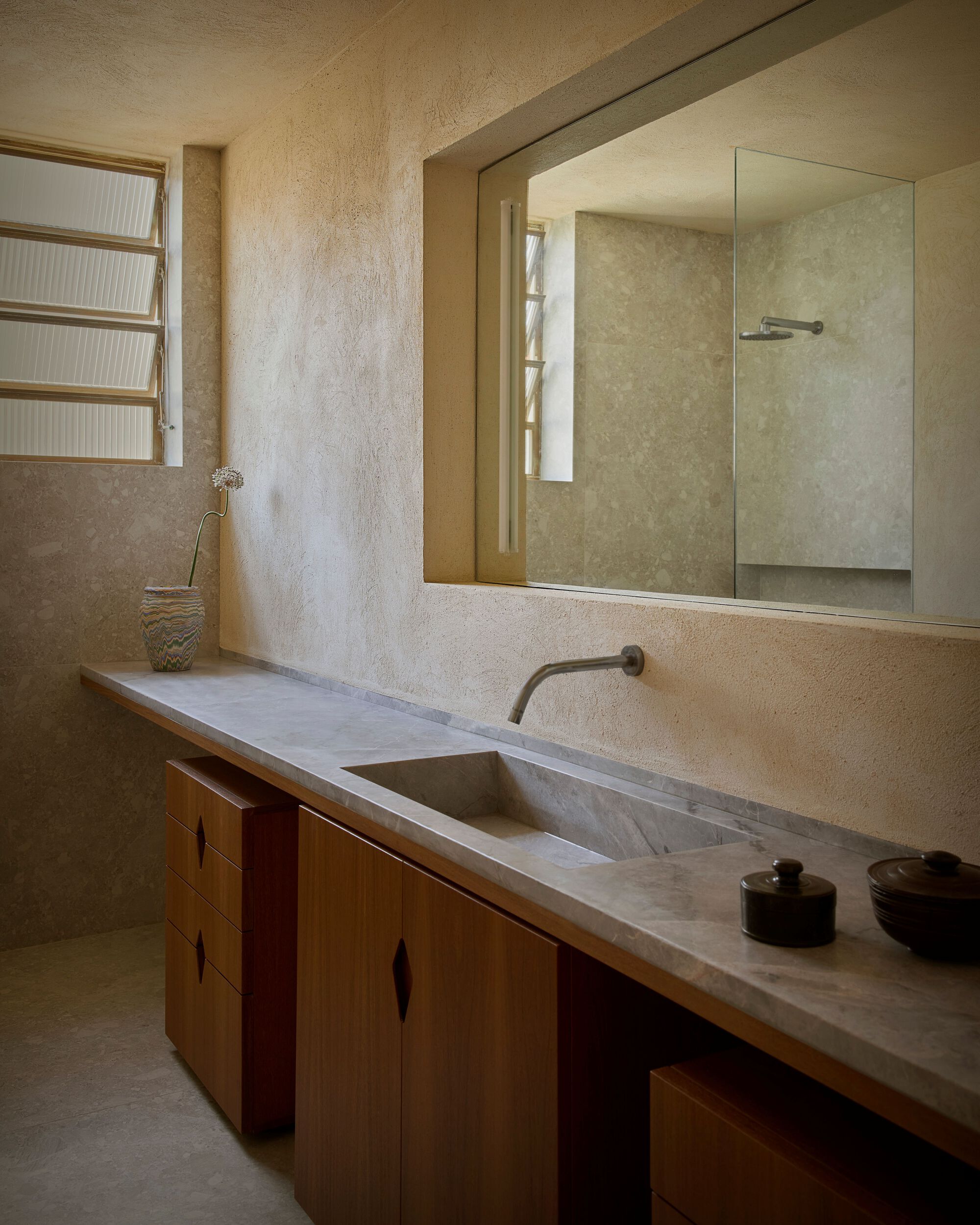 Bathroom with a long stone vanity, integrated sink and timber cabinets under a wide mirror, with a louvred window and dried flowers
