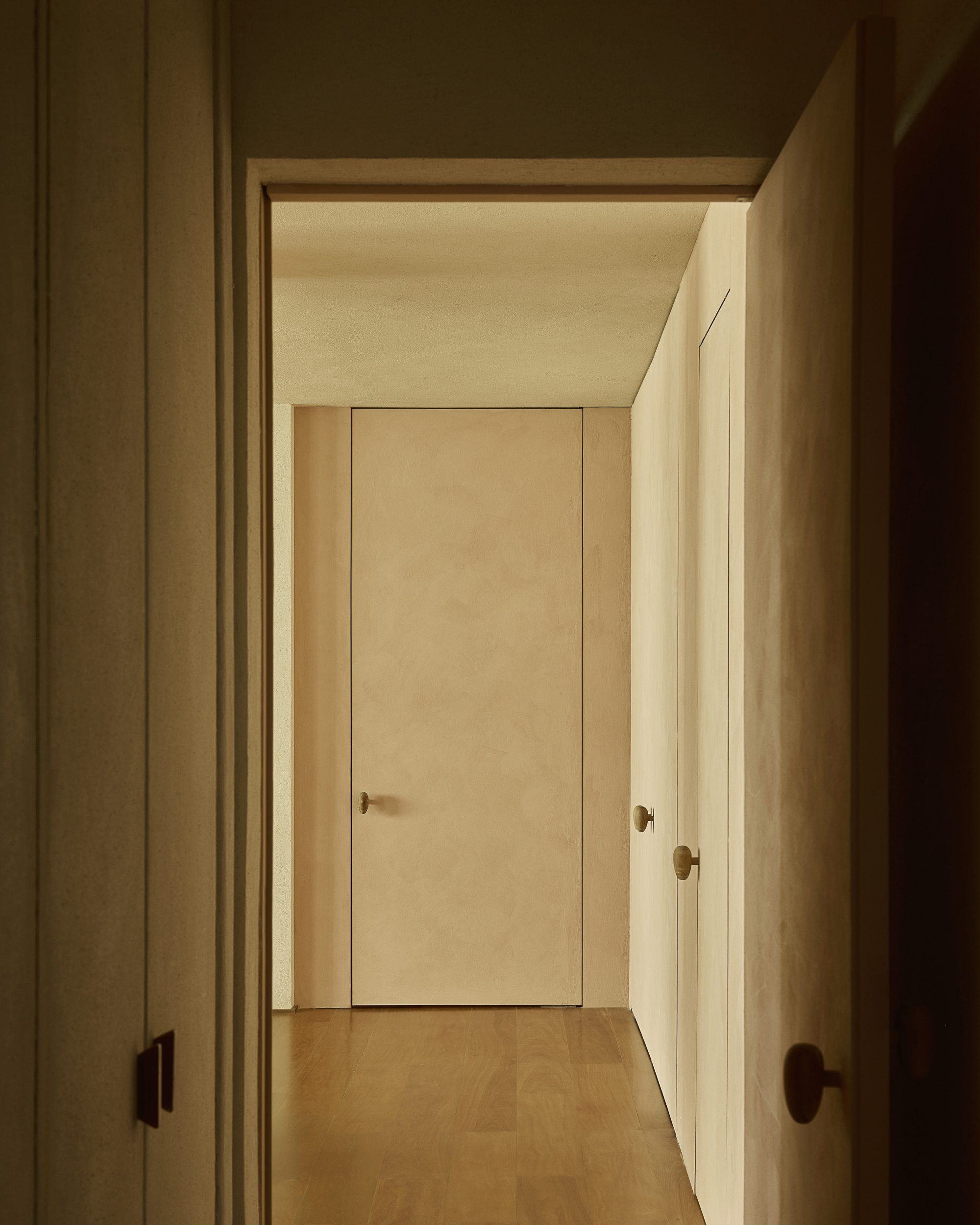 Narrow hallway with flush plaster doors and round brass knobs, lit by soft low daylight on a timber floor
