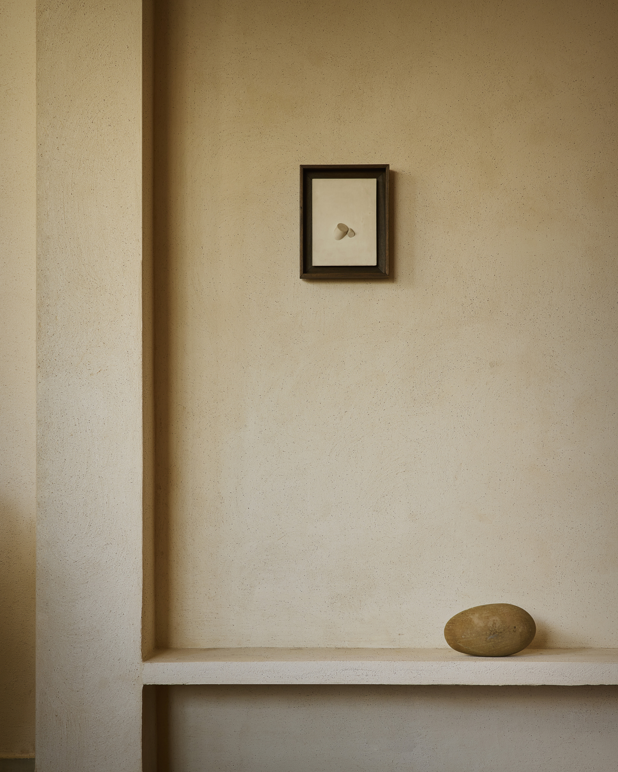 Framed small artwork above a plaster shelf holding a single smooth stone, in warm raking light