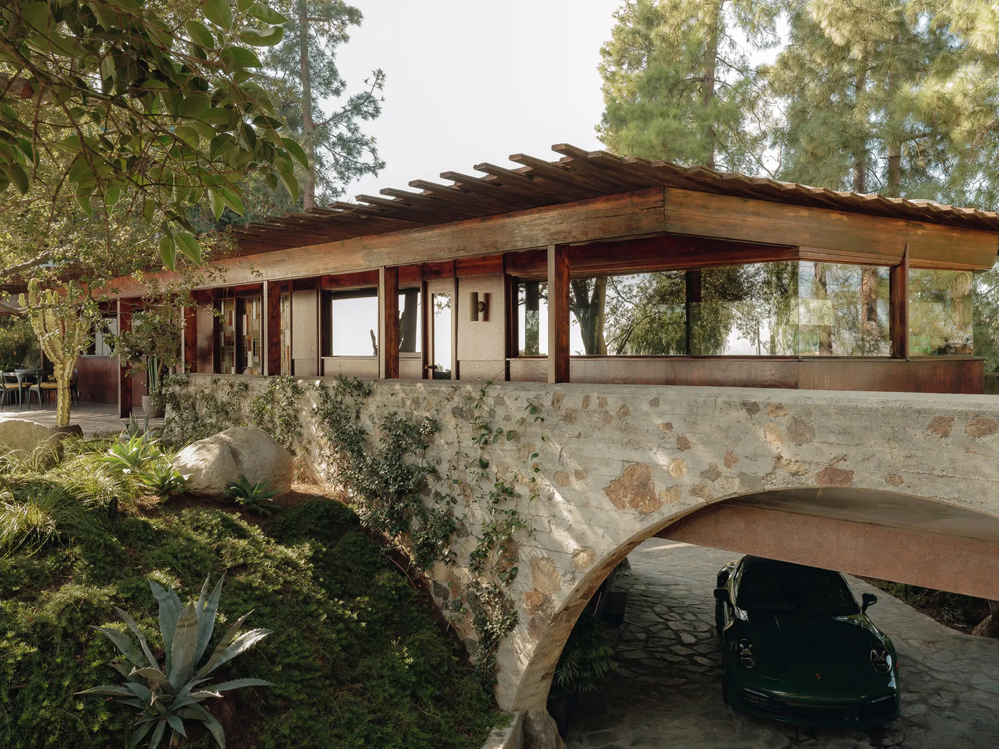Timber pavilion with pergola roof resting on a rough stone arch, a dark green car parked beneath in dappled light