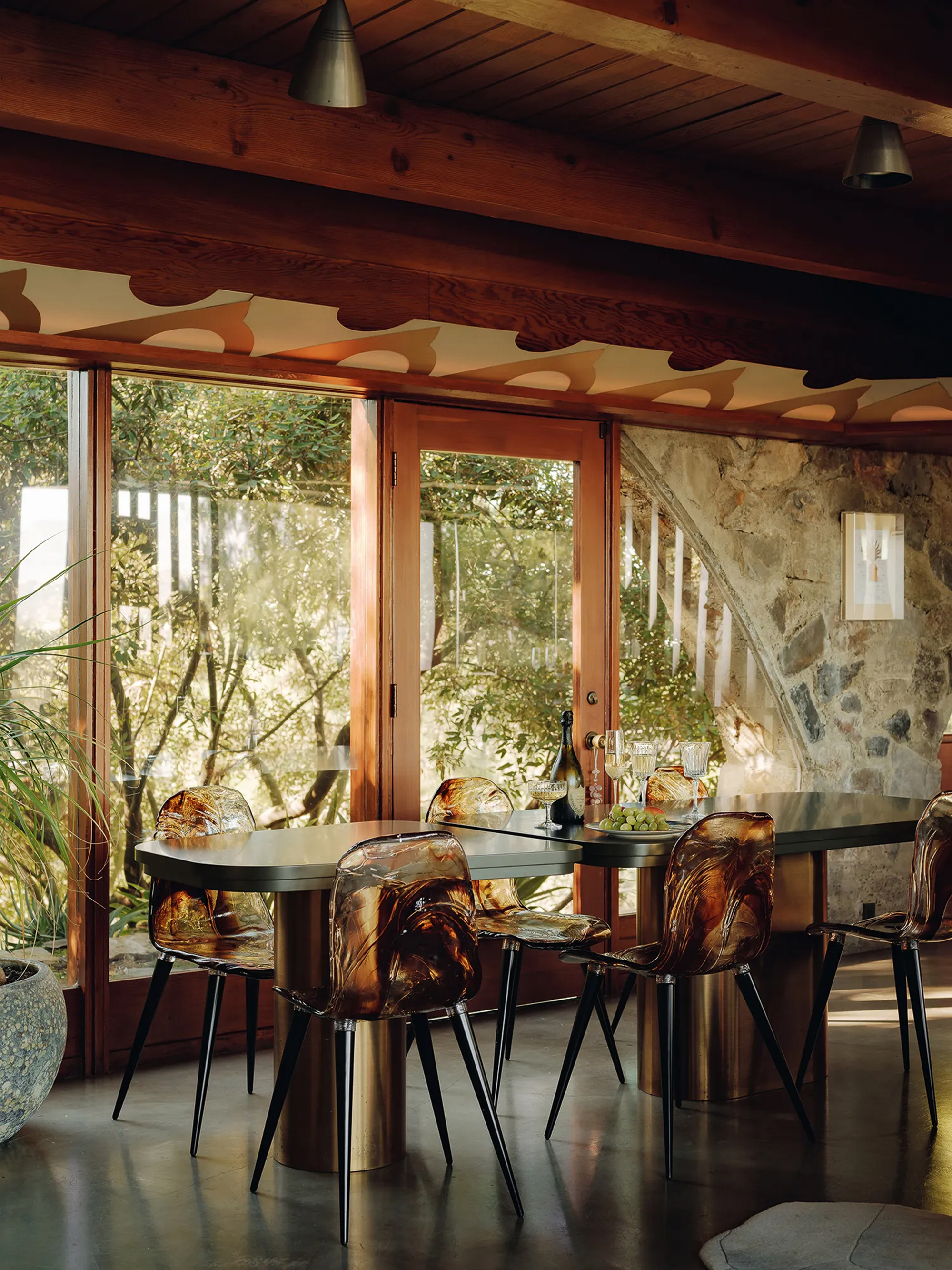 Dining table with amber translucent chairs beside floor-to-ceiling glazing and a rough stone wall under exposed wood beams