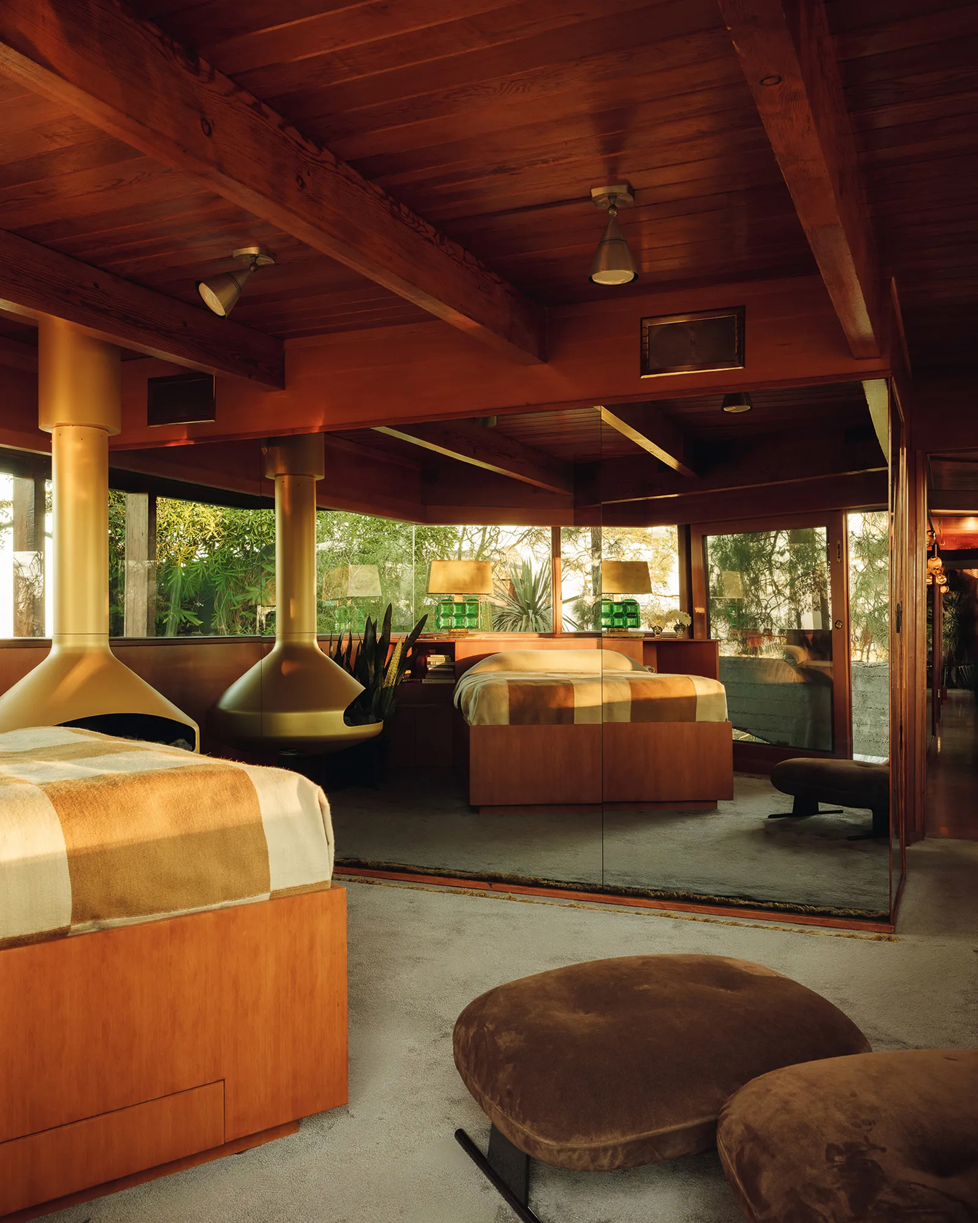 Bedroom with two suspended brass fireplaces, wood-paneled ceiling and a mirrored wall reflecting the beds in warm evening light