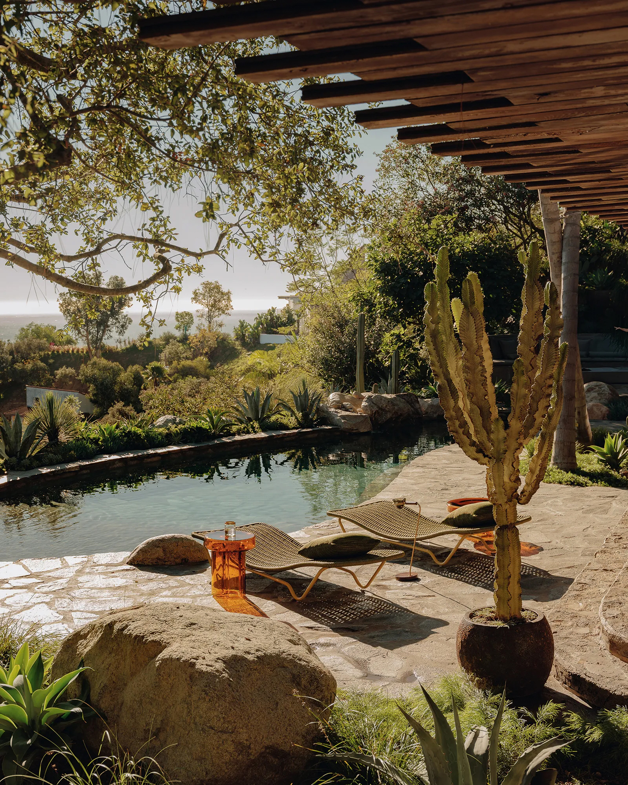 Woven loungers and an amber side table on flagstone paving beside a pool, with a tall cactus and ocean haze beyond