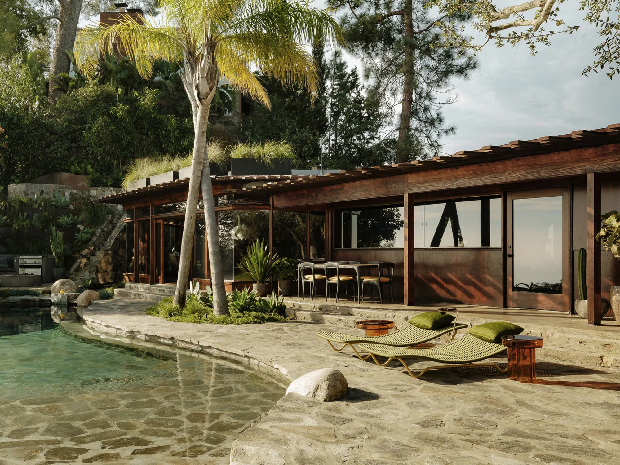 Pool edge of flagstone and boulders in front of a long timber pavilion, with a leaning palm and green loungers