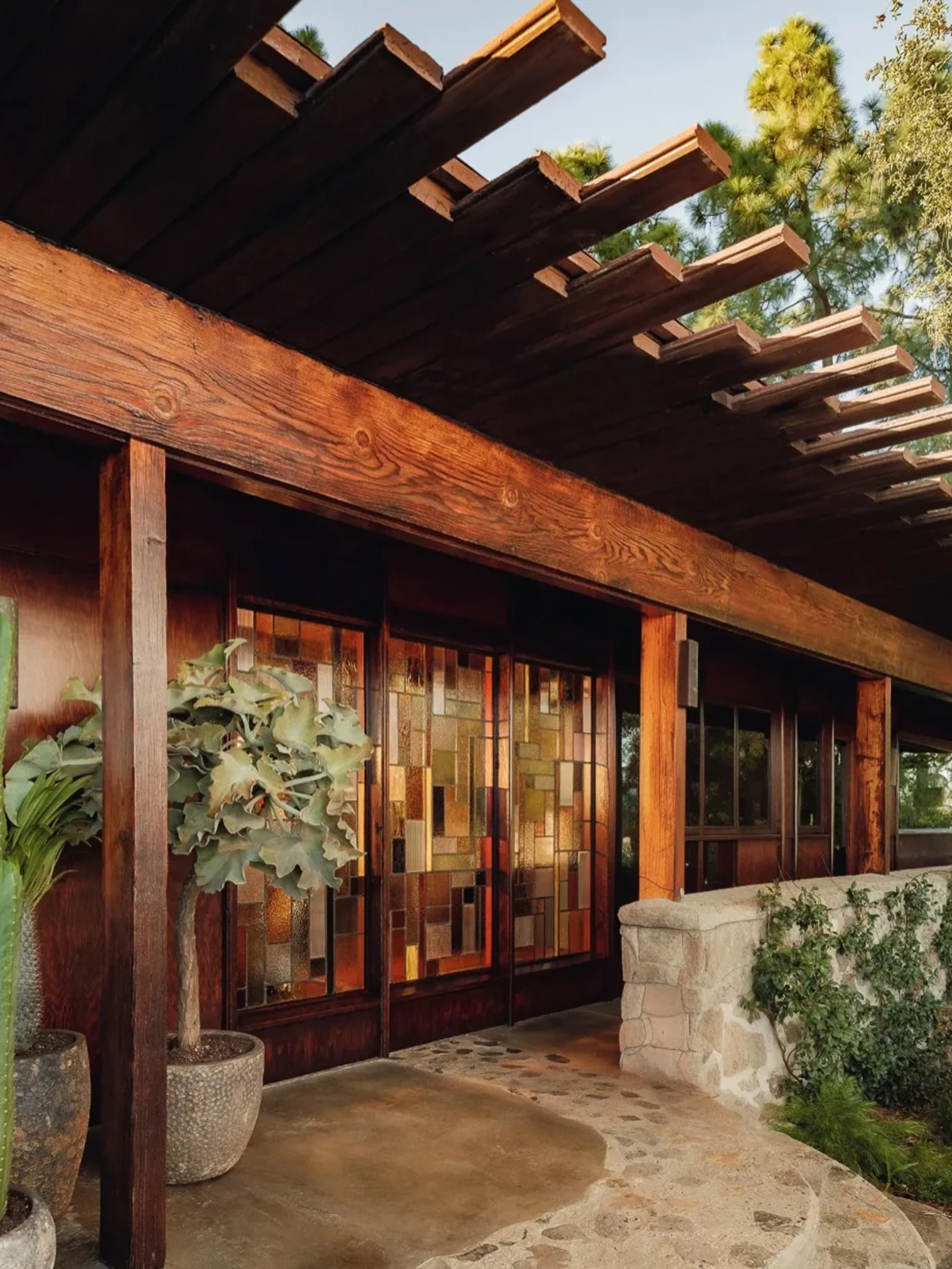 Timber porch with stepped rafters and stained glass panel doors above a flagstone path and a potted silver-leaf plant in afternoon light