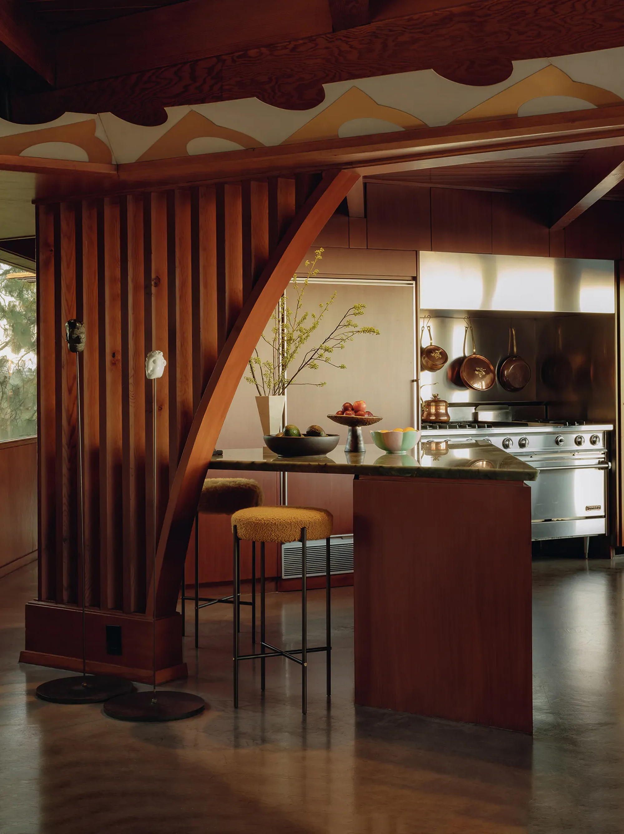 Kitchen with curved timber brace, slatted wood screen, stone-topped island and copper pans hanging above a steel range in warm light