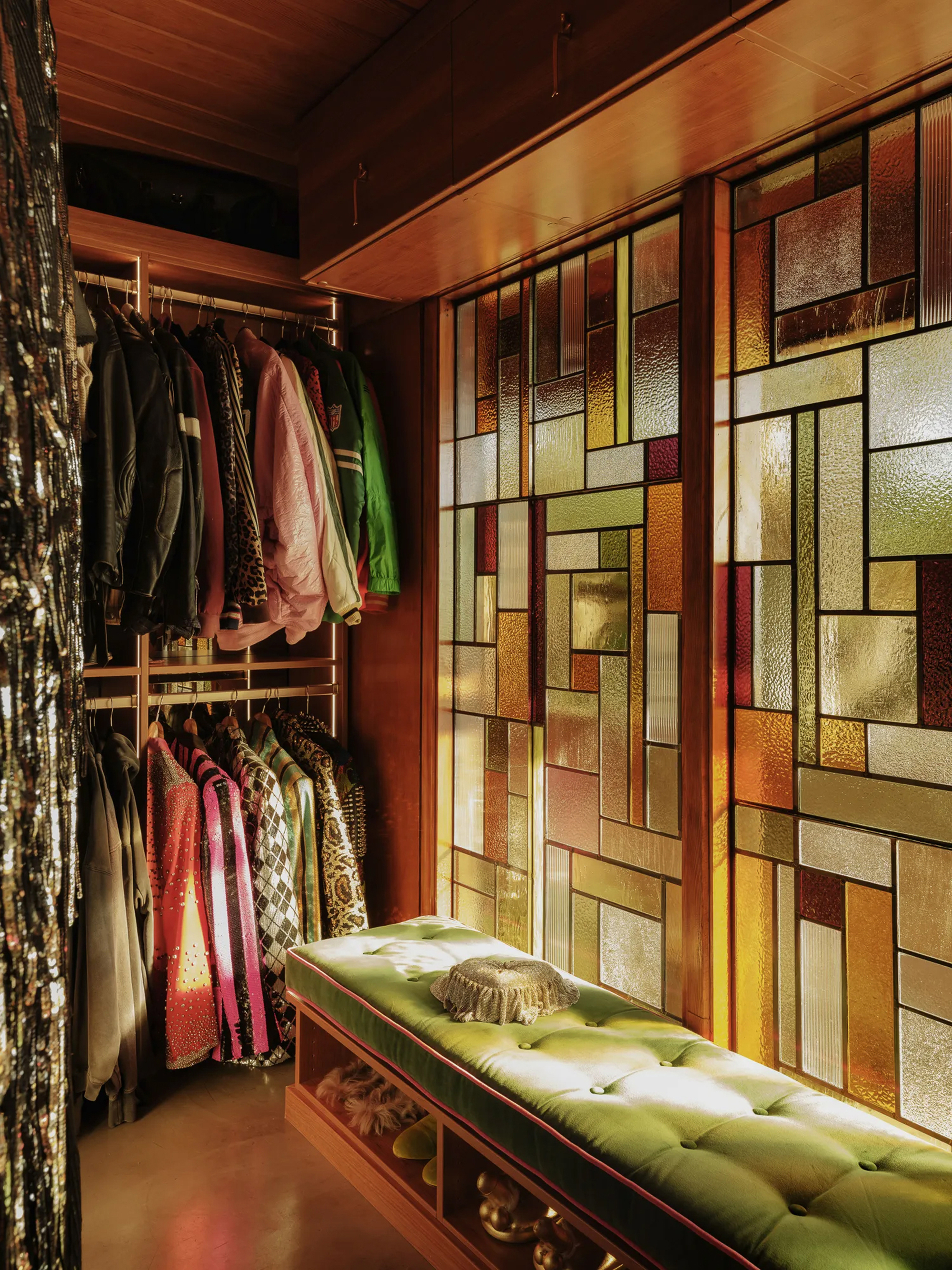 Dressing room with colored glass panel wall, green tufted velvet bench and hanging jackets, lit by a warm glow from behind the glass