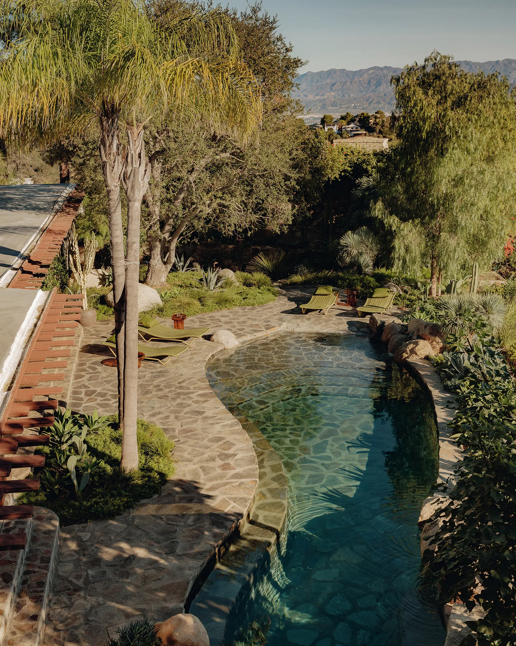 Raised view of a kidney-shaped pool edged in flagstone with lounge chairs, palms and oaks, and distant hills in clear daylight